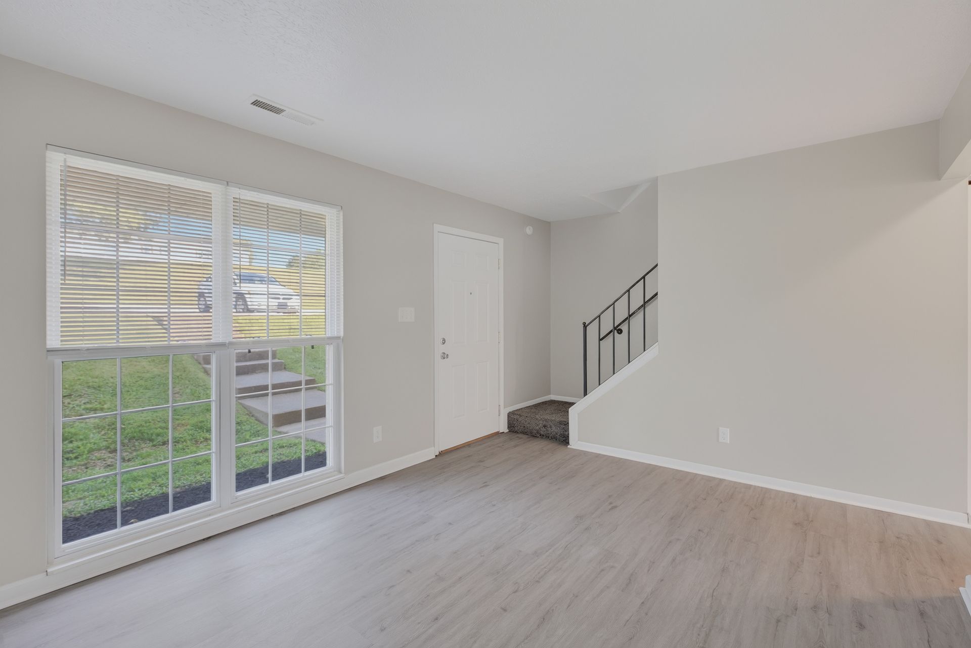 Empty living room with white walls, sliding glass doors, light wood floor, and stairs at the back.