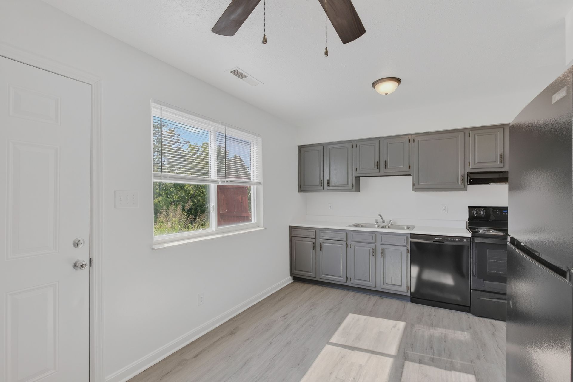 Empty kitchen with gray cabinets, black appliances, white walls, and a window letting in sunlight.