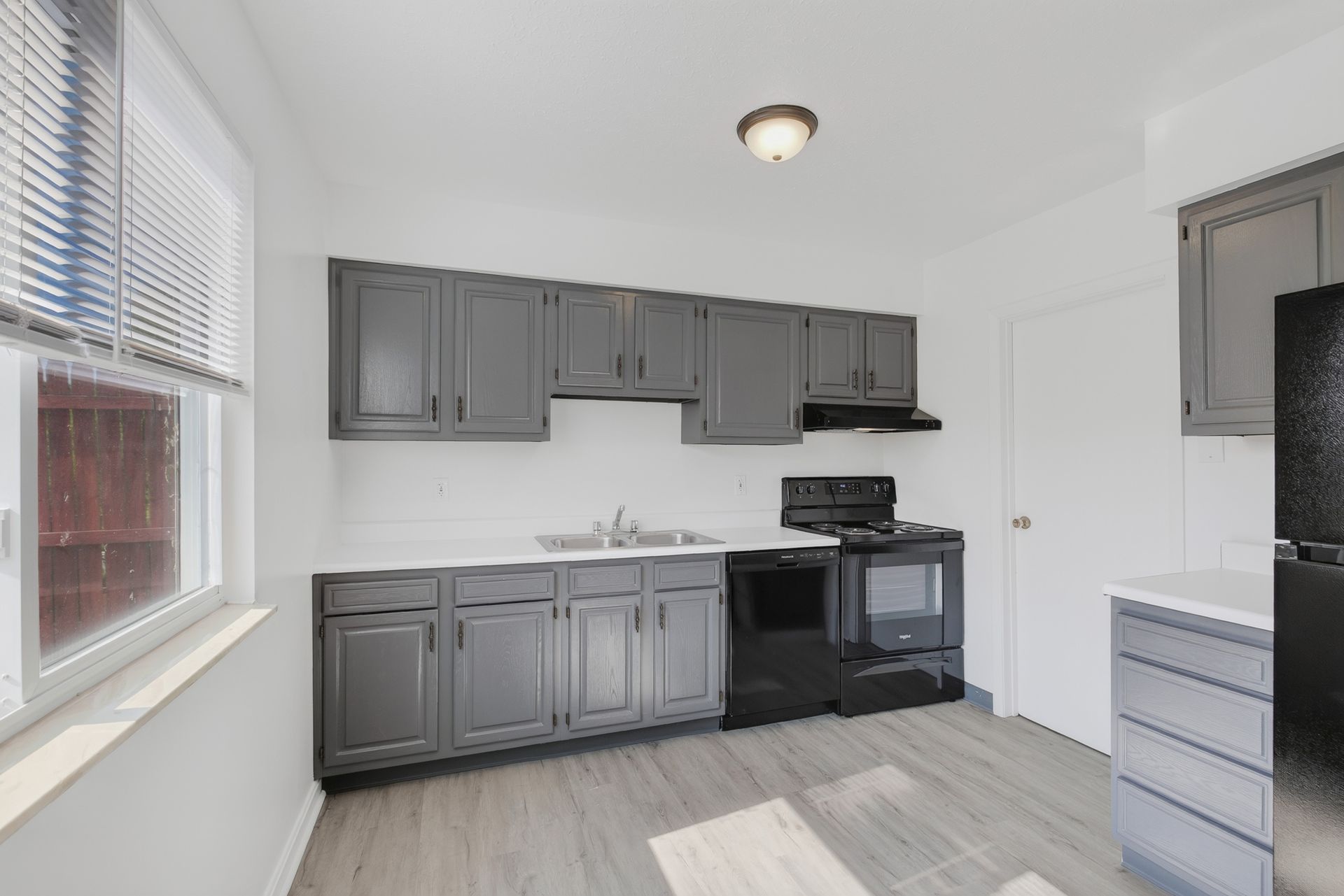 Empty kitchen with gray cabinets, black stove, white walls, and a window with blinds.