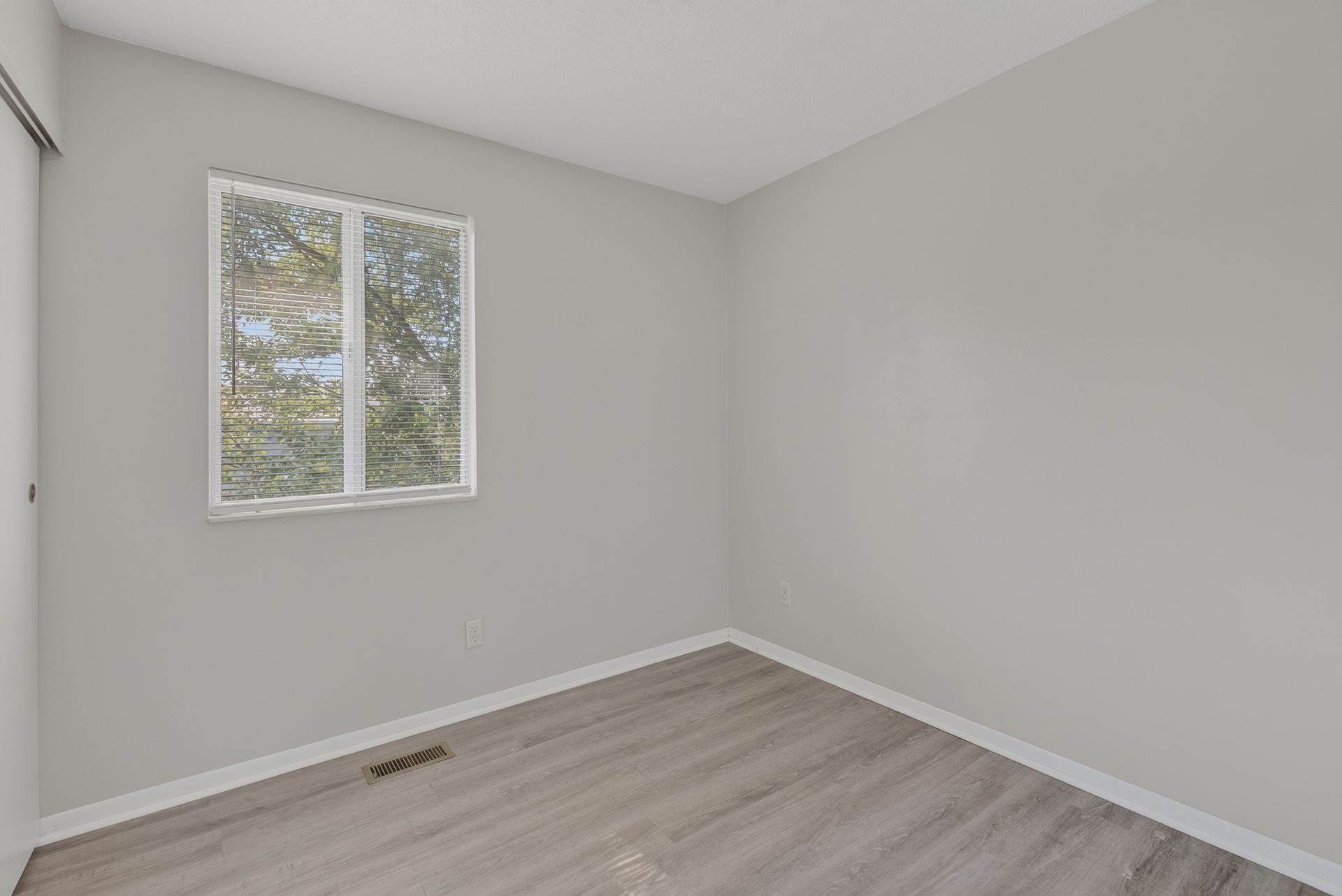 Empty gray bedroom with light wood flooring and a window on the left wall