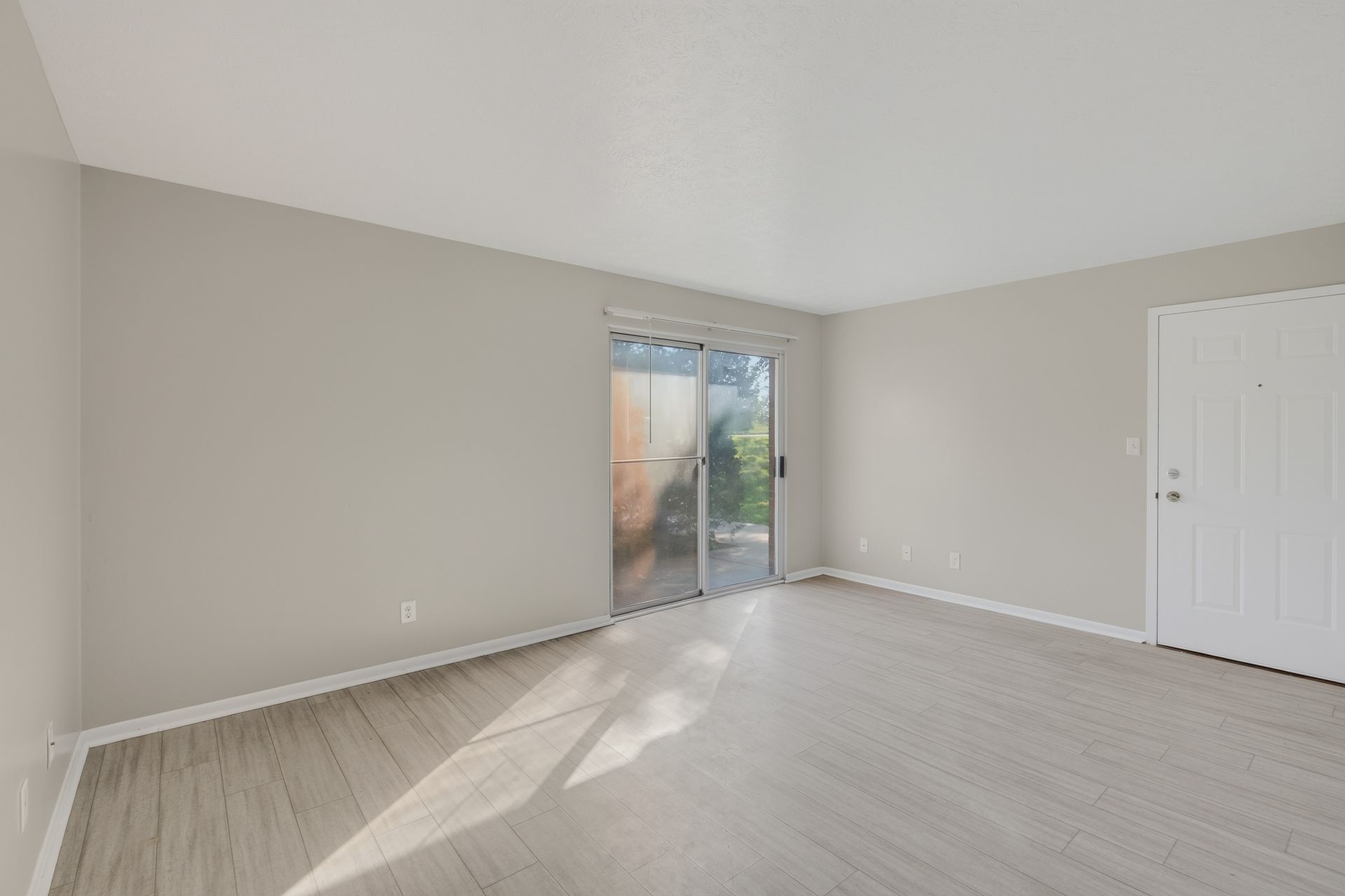 Empty beige bedroom with white closet doors and sliding glass door to a patio, bright sunlight on carpeted floor