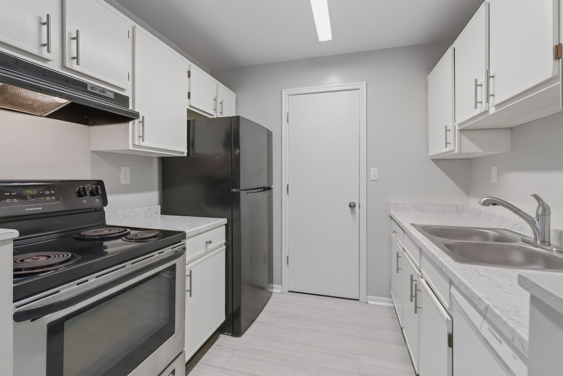 Narrow galley kitchen with white cabinets, black appliances, and a door at the far end.