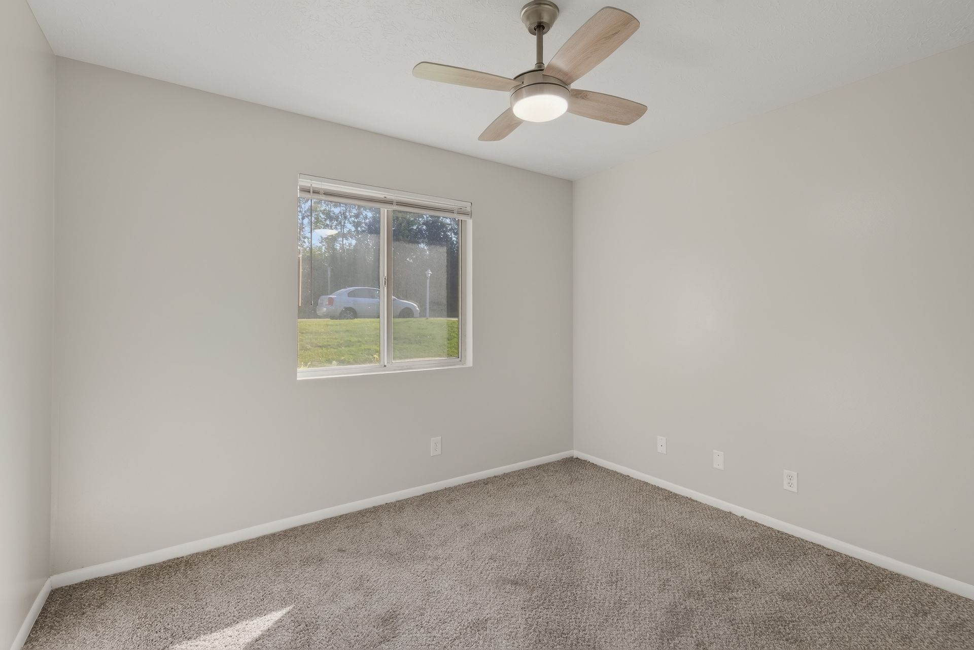 Empty gray bedroom with carpet, ceiling fan, and a window overlooking greenery.