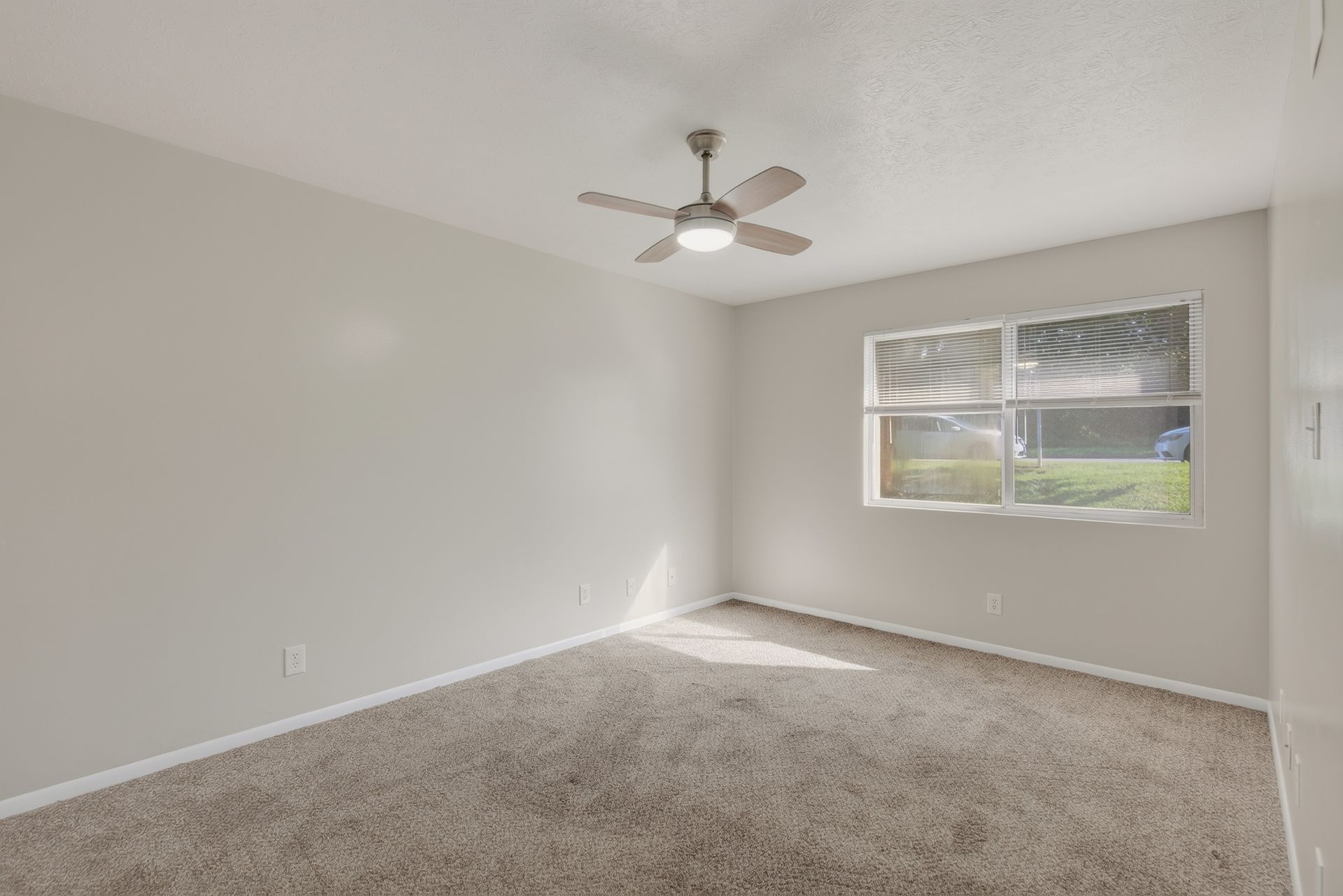 Empty beige bedroom with carpet, white walls, ceiling fan, and a window letting in daylight