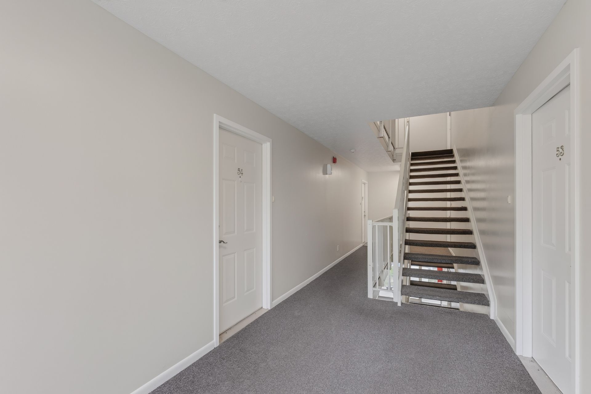 Empty beige hallway with carpet, white doors, and stairs leading up to a loft area.