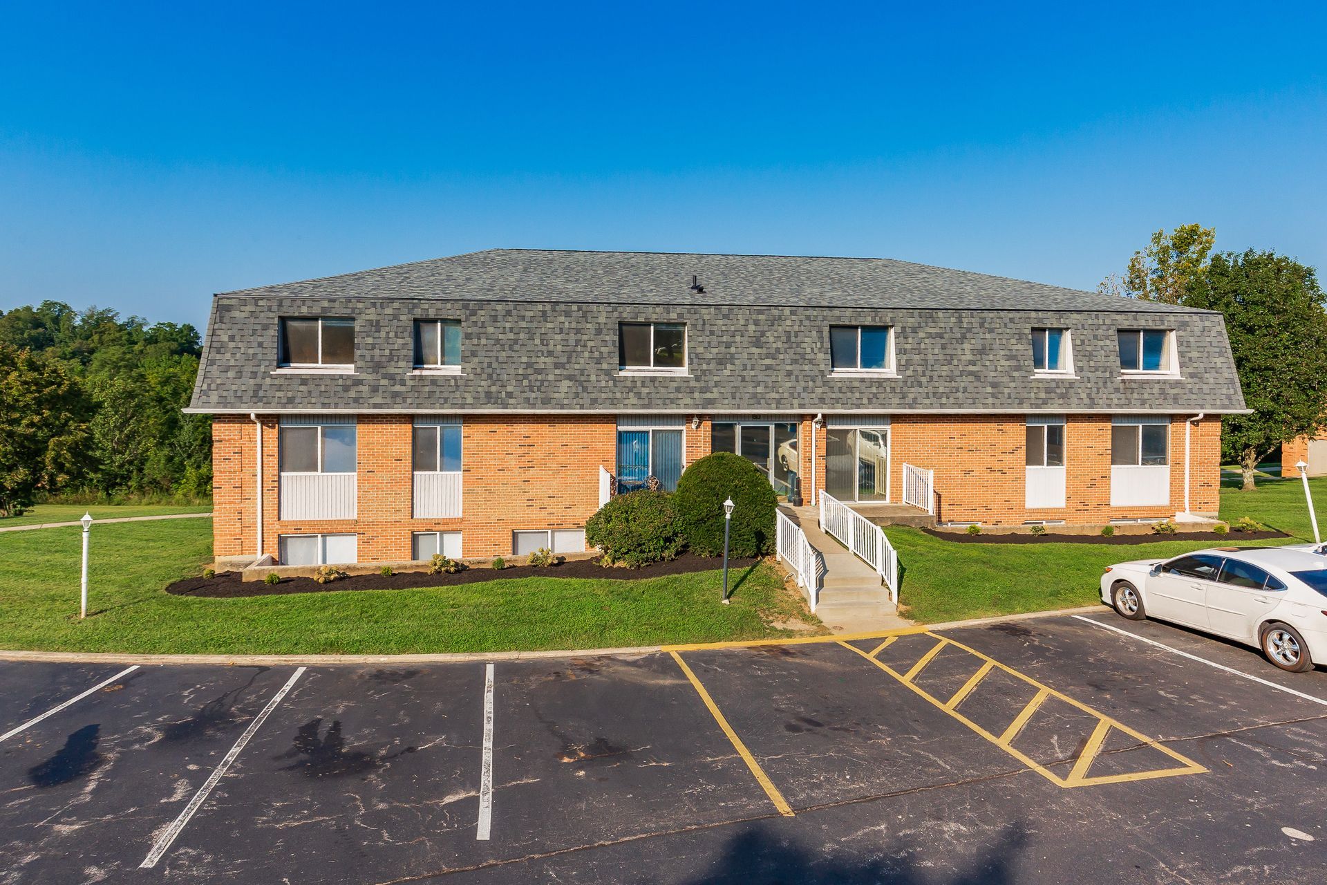 Two-story brick apartment building with dark roof, front steps, and a parking lot under a clear blue sky