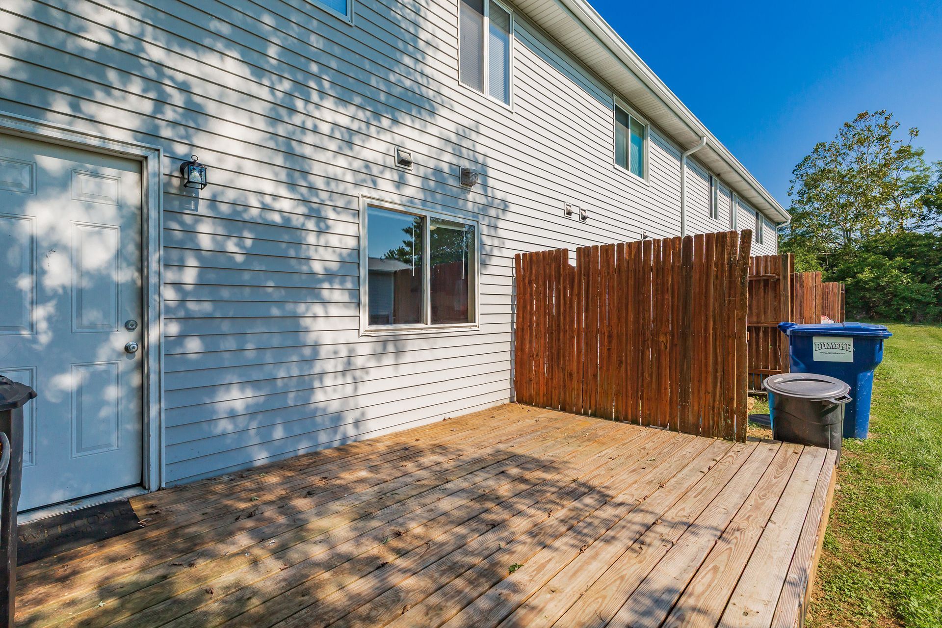 Wooden backyard deck beside white siding, with a fence, windows, and two trash bins.