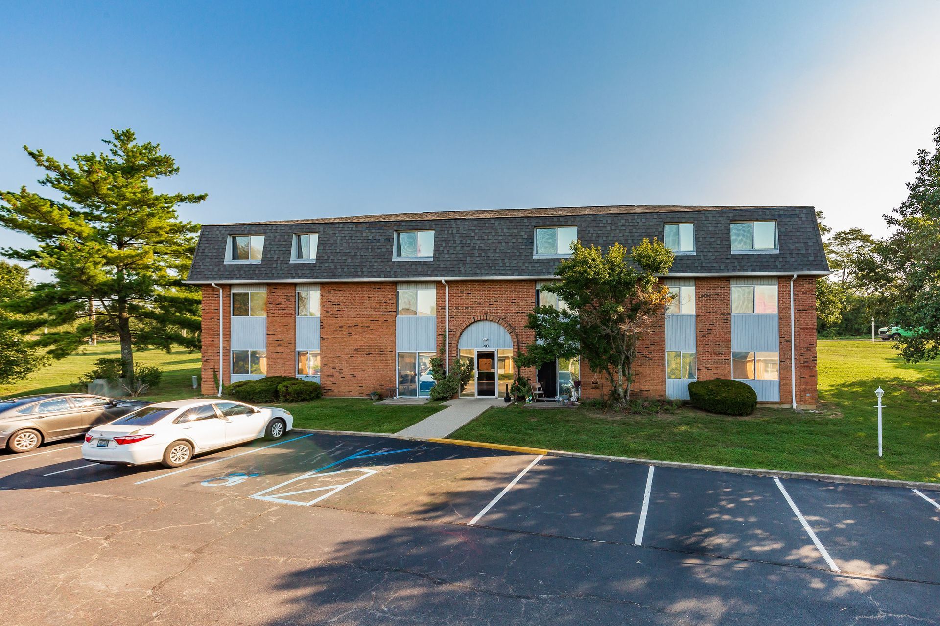 Brick apartment building with parked cars and trees in a sunny lot