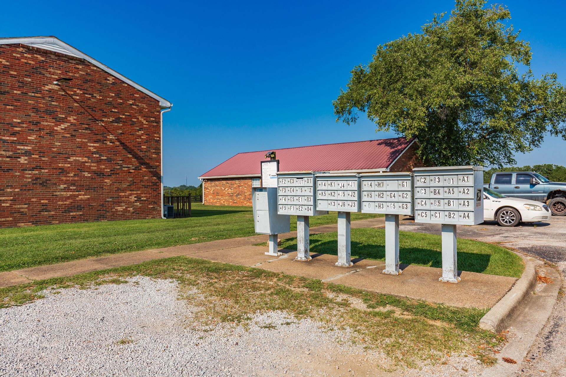 Mailboxes on posts beside a gravel lot, with a brick building, tree, and parked car under a blue sky