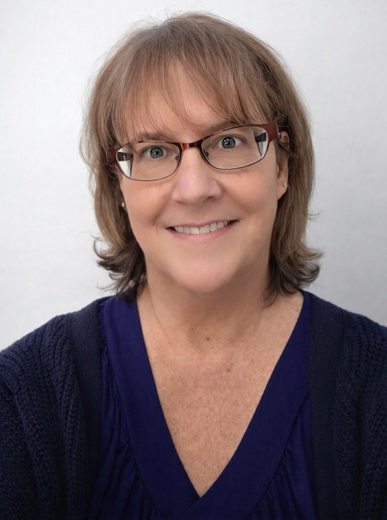 Smiling person with glasses and shoulder-length brown hair wearing a navy top against a light background