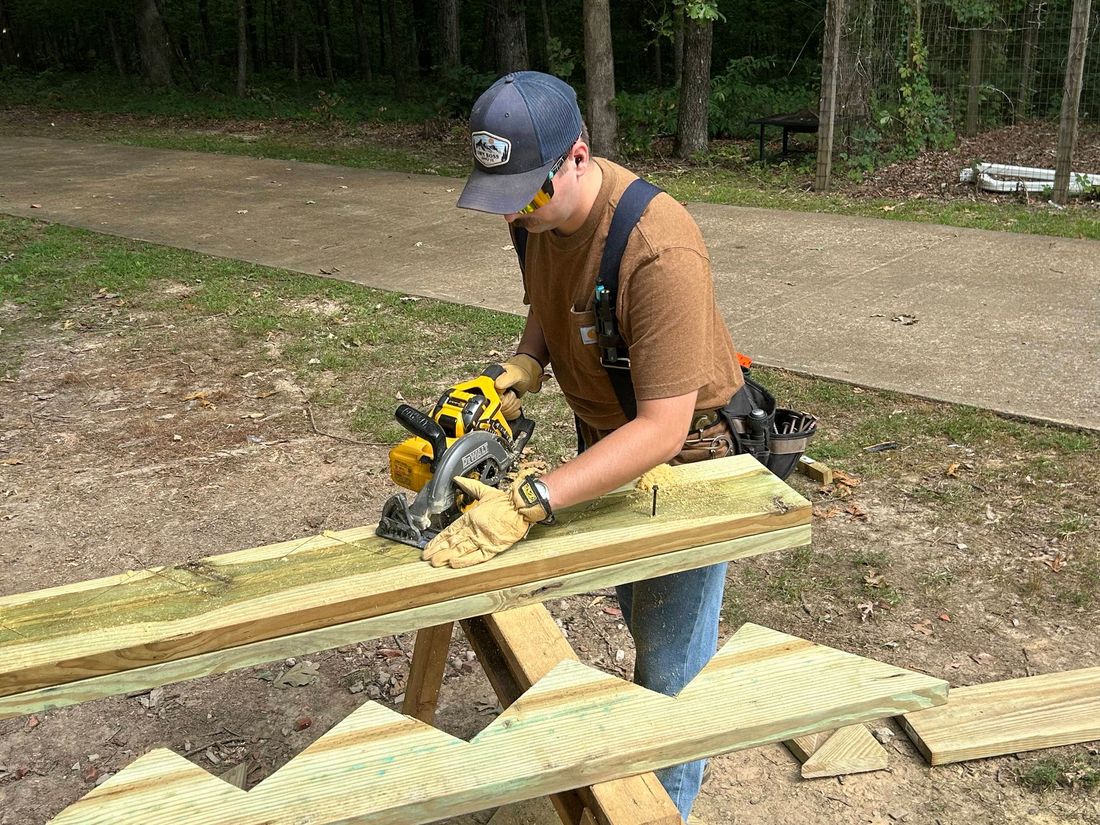 Person using a circular saw to cut a wooden plank outdoors. Wears safety glasses, gloves, and a tool belt.