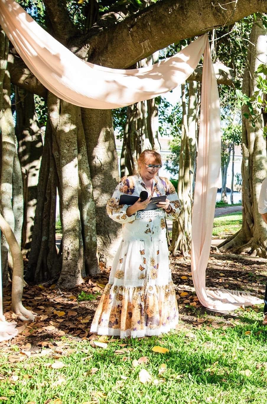A female marriage celebrant in a white floral dress is standing under a tree holding a microphone.
