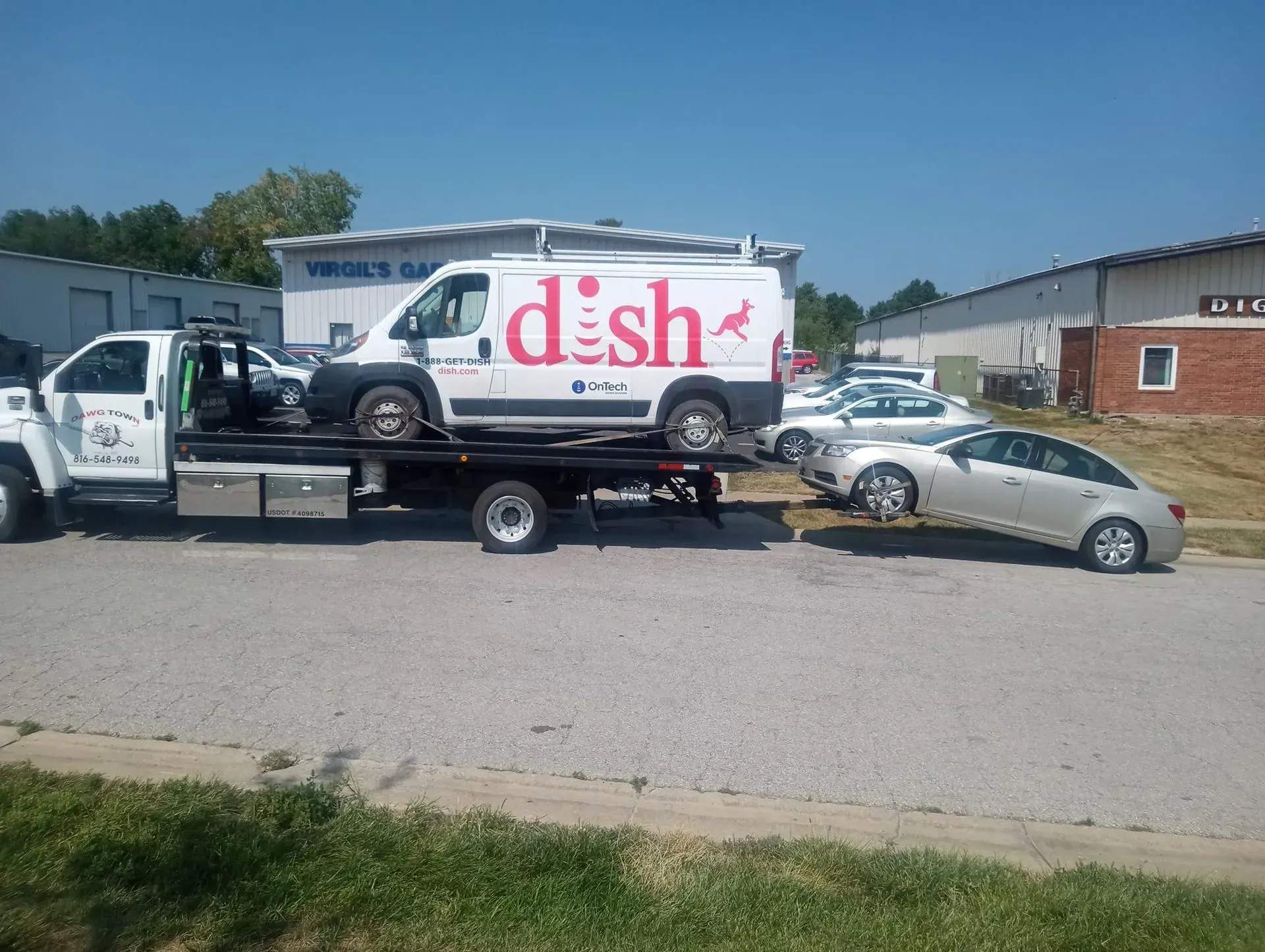 Tow truck hauling a Dish-branded SUV, with a damaged silver car on a street near buildings.