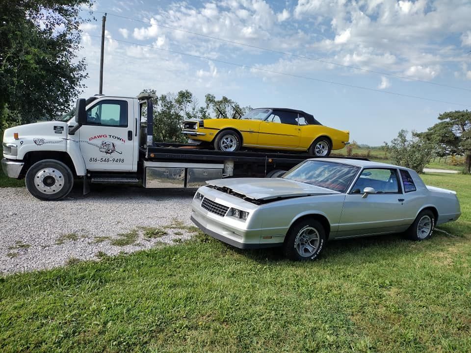 Tow truck carrying a yellow sports car, with a silver coupe parked on grass in front