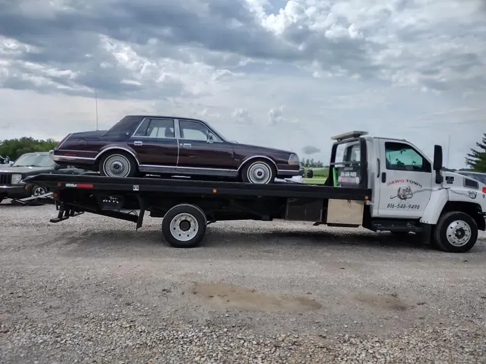 Tow truck carrying a maroon sedan on a flatbed under a cloudy sky.