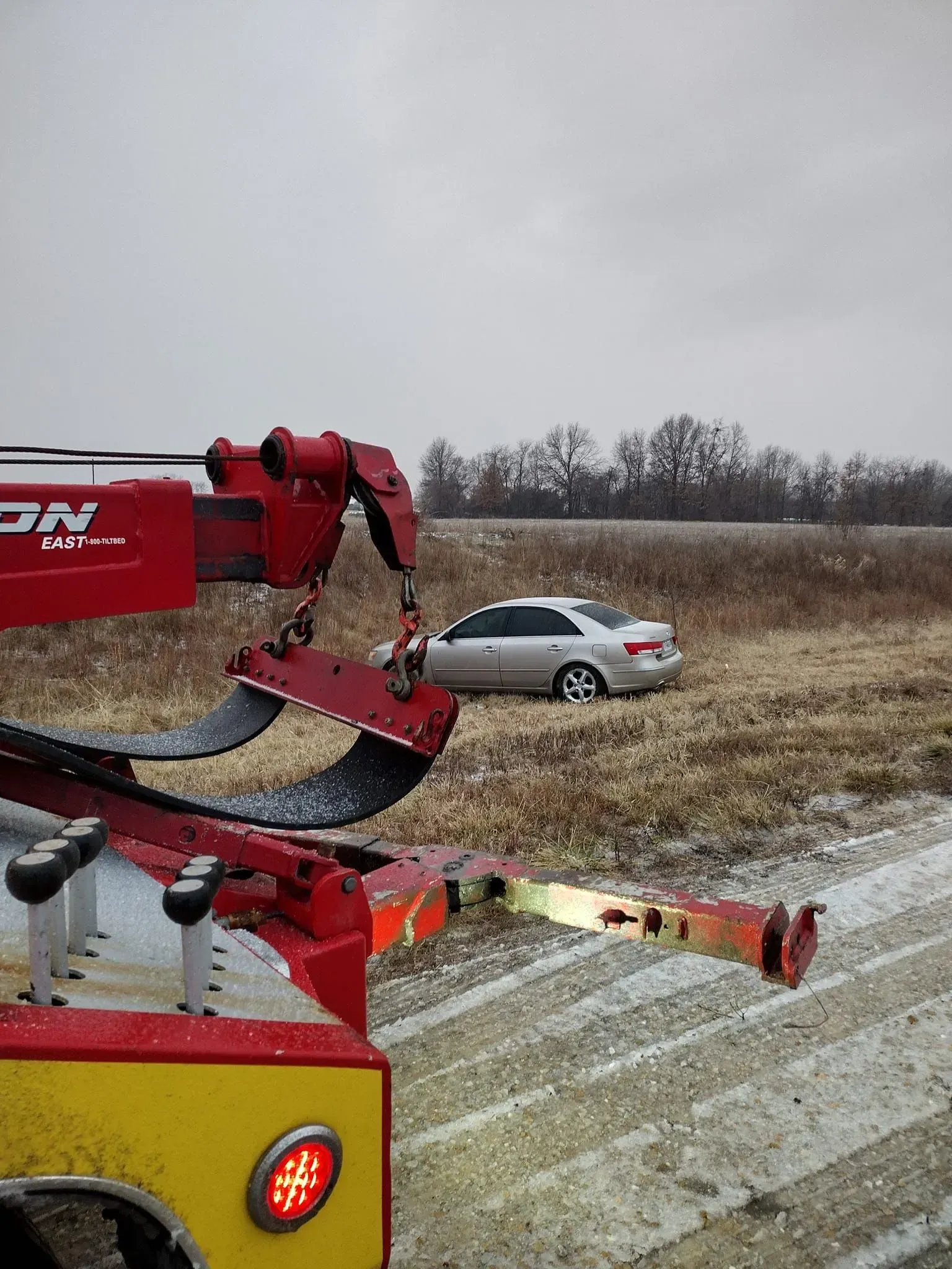 Tow truck lifting a silver sedan on a snowy roadside, with red equipment in the foreground