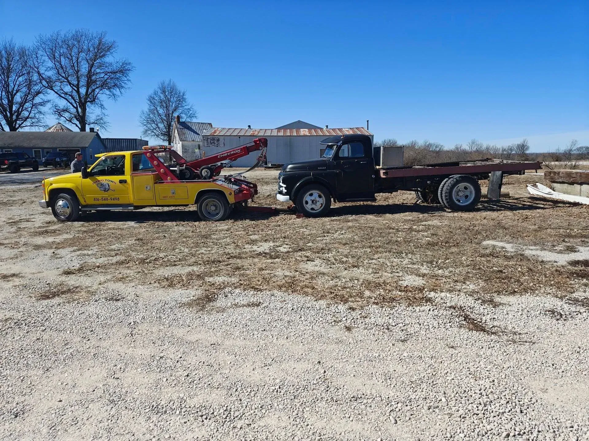 Two tow trucks on a gravel lot under a clear blue sky, one yellow and one black.