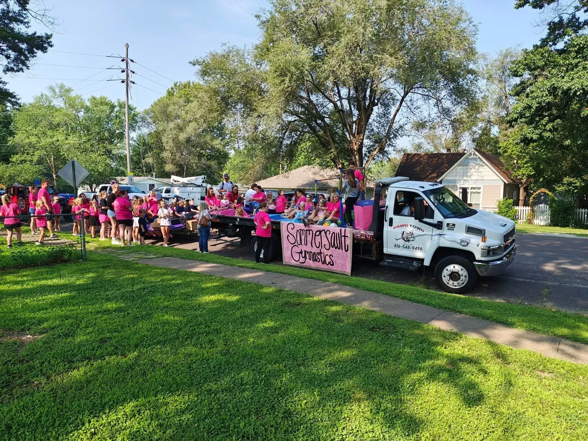 Pink-clad crowd marching beside a white truck in a sunny neighborhood parade
