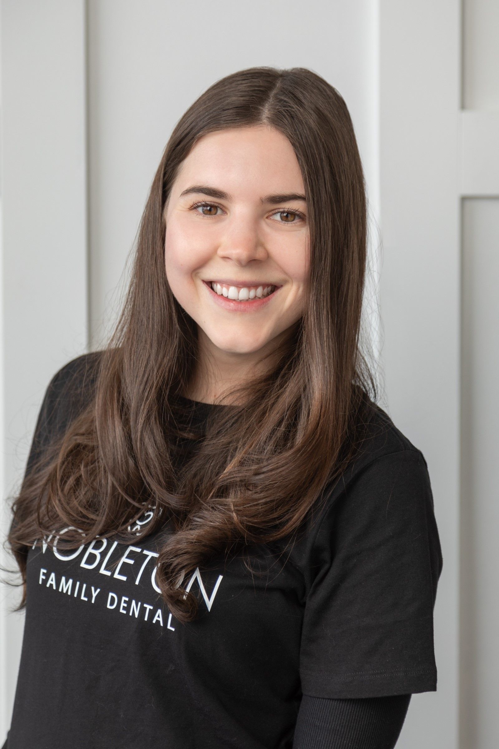 Woman with brown hair smiling, wearing a black shirt, in front of a white wall.