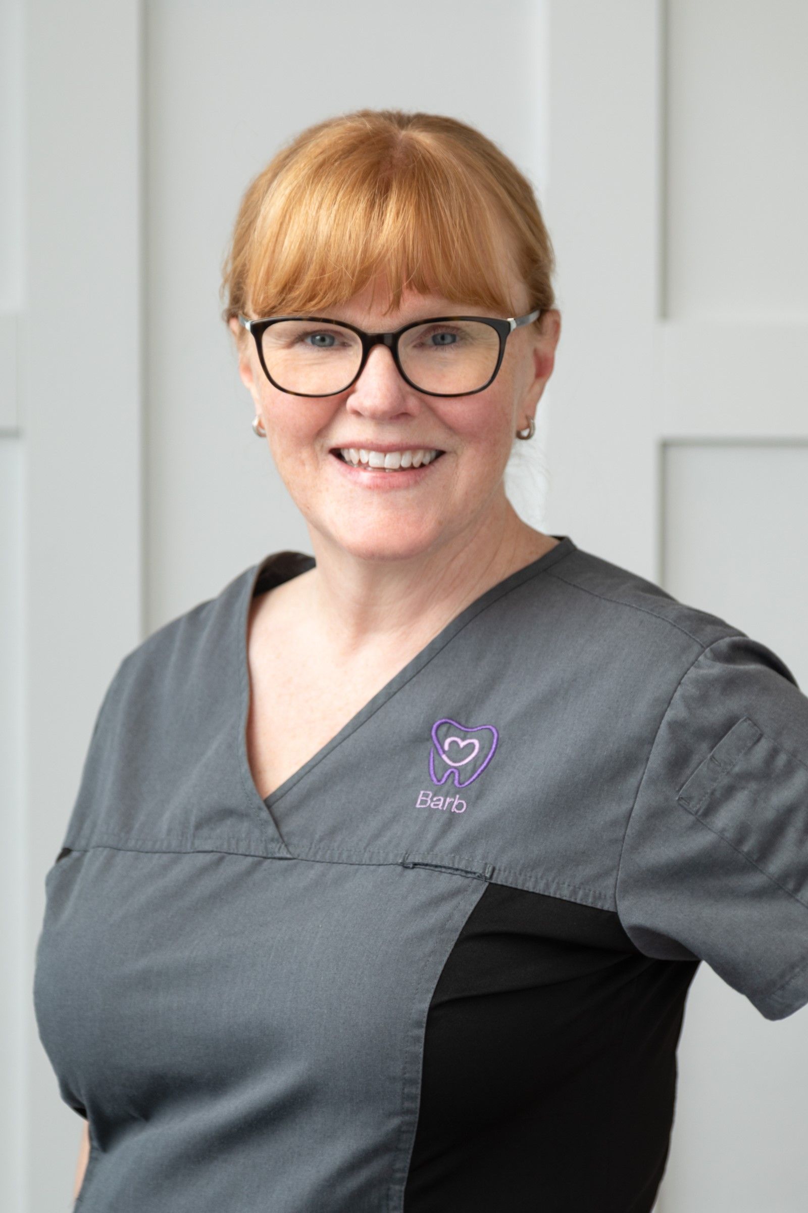 Woman in gray scrubs with glasses smiling, in a clinic setting.