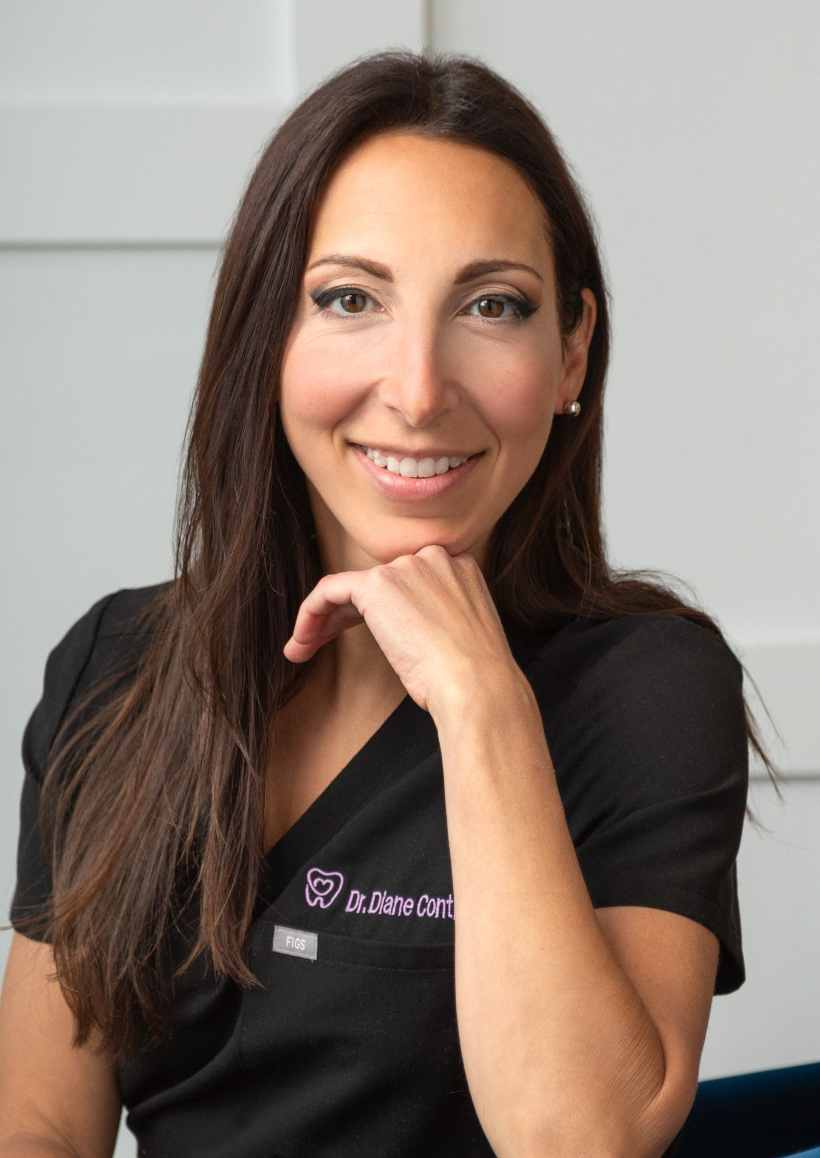 Woman with long brown hair, smiling, hand on chin, wearing black scrubs.