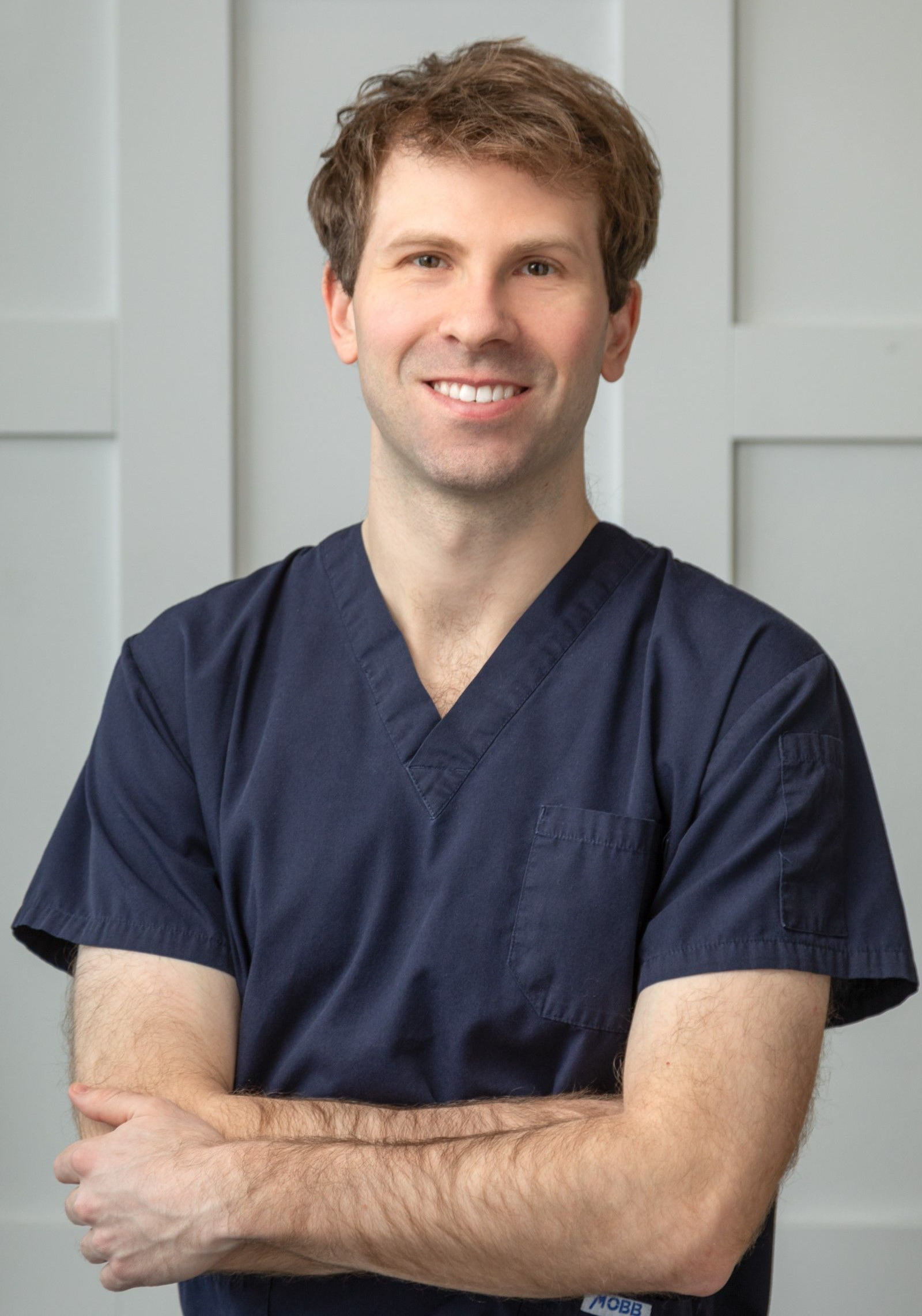 Man in navy scrubs with arms crossed, smiling in front of a white paneled wall.