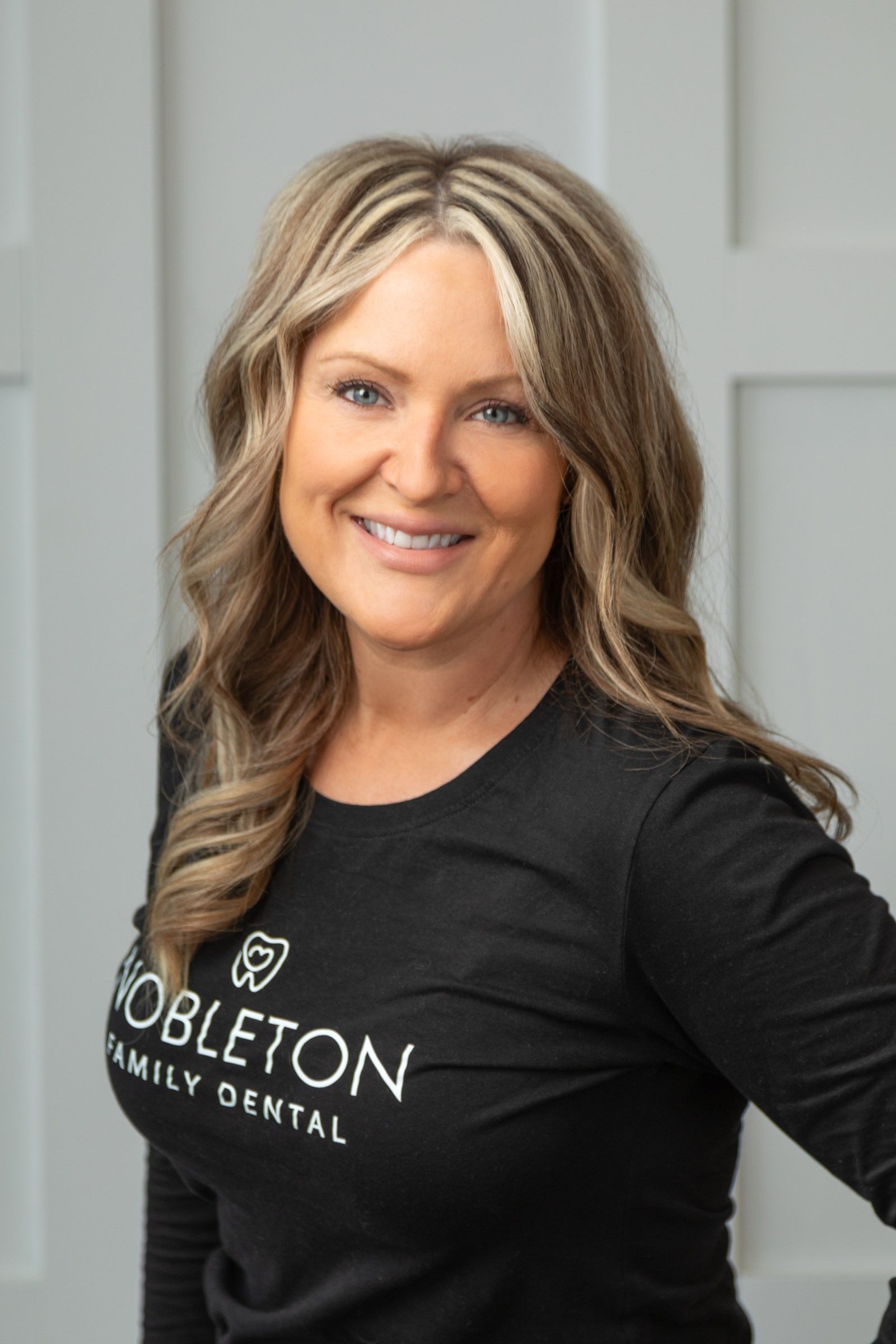 Woman with blonde highlights smiles in front of a wall, wearing a black