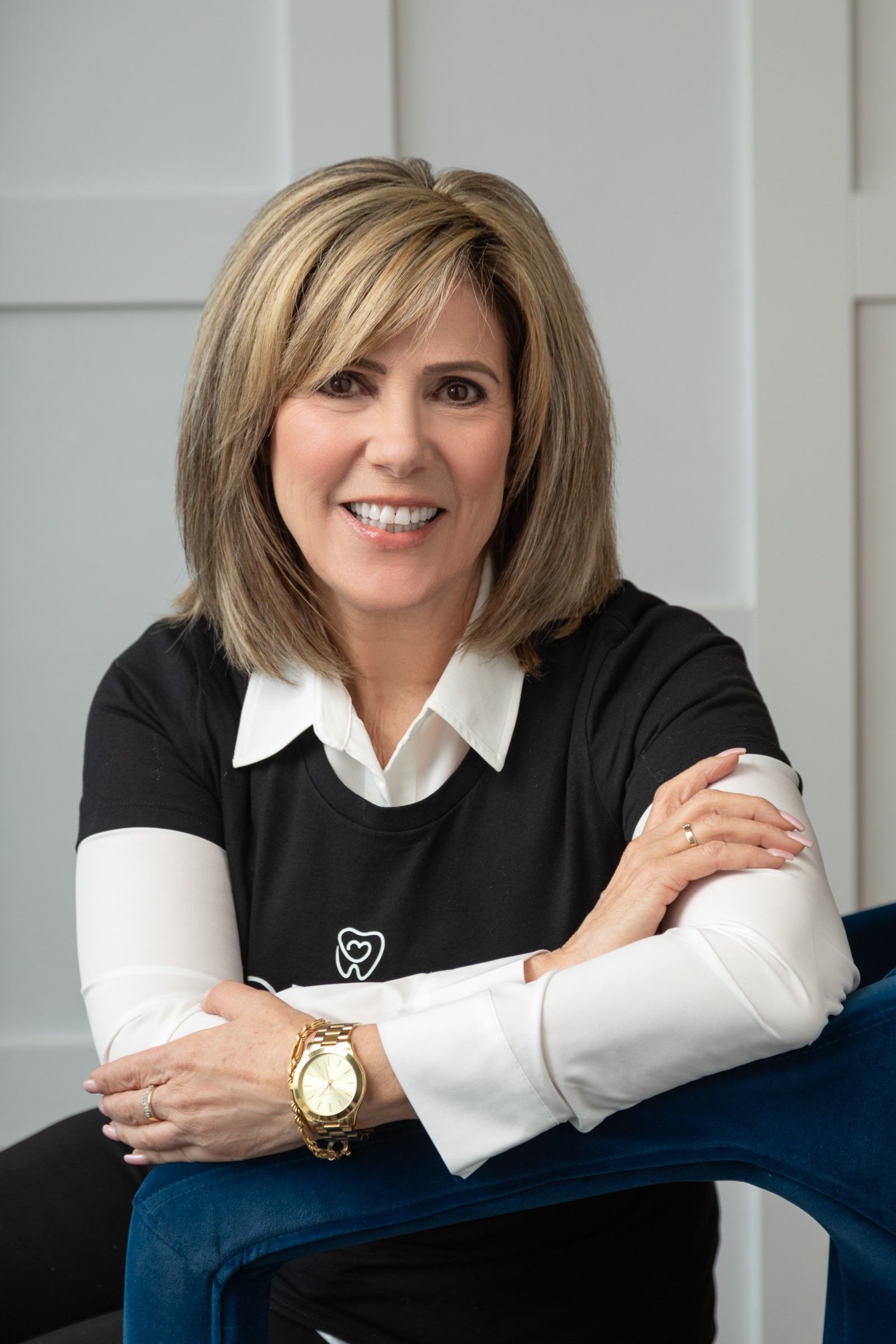 Woman with blonde hair, smiling, arms crossed, seated in blue chair, wearing black shirt, white undershirt, and gold watch.