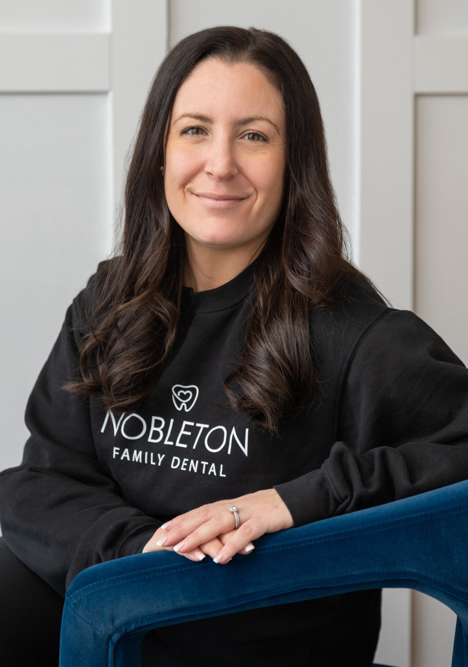 Woman with dark hair in a black sweatshirt for Nobleton Family Dental smiles, seated in front of a white wall.
