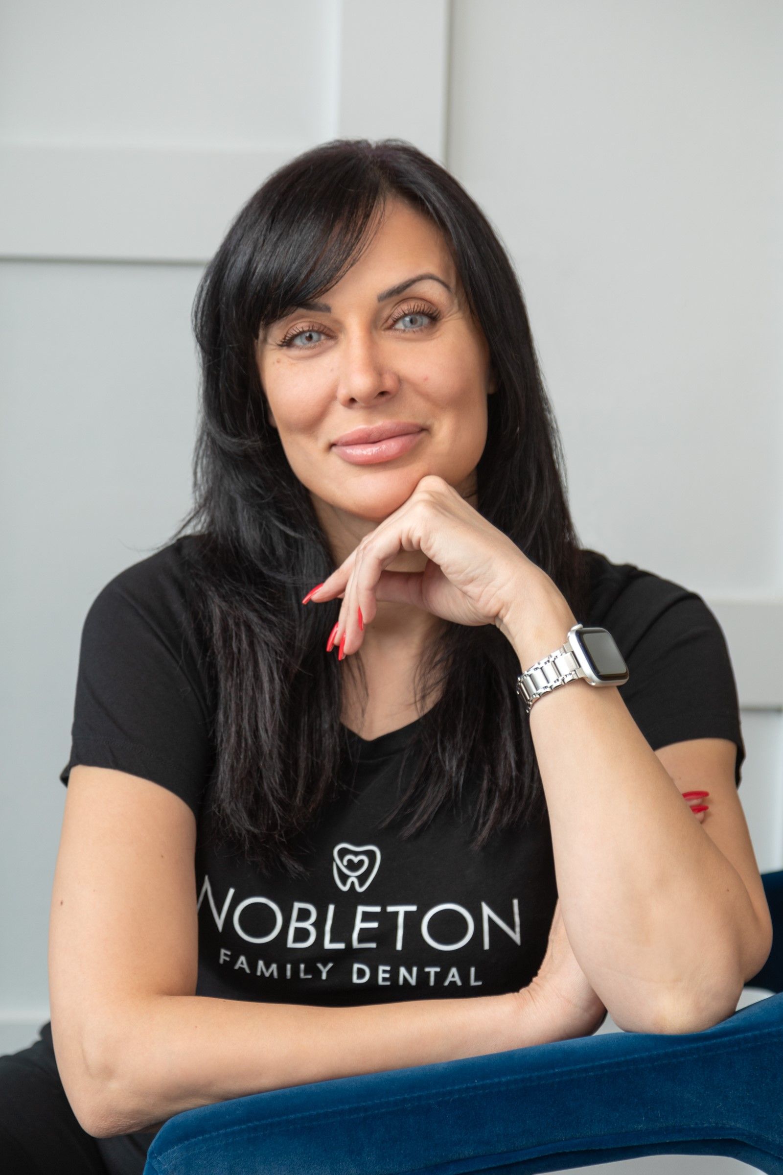 Woman with dark hair, wearing a black t-shirt, seated, smiling, hand on chin, in a dental office.