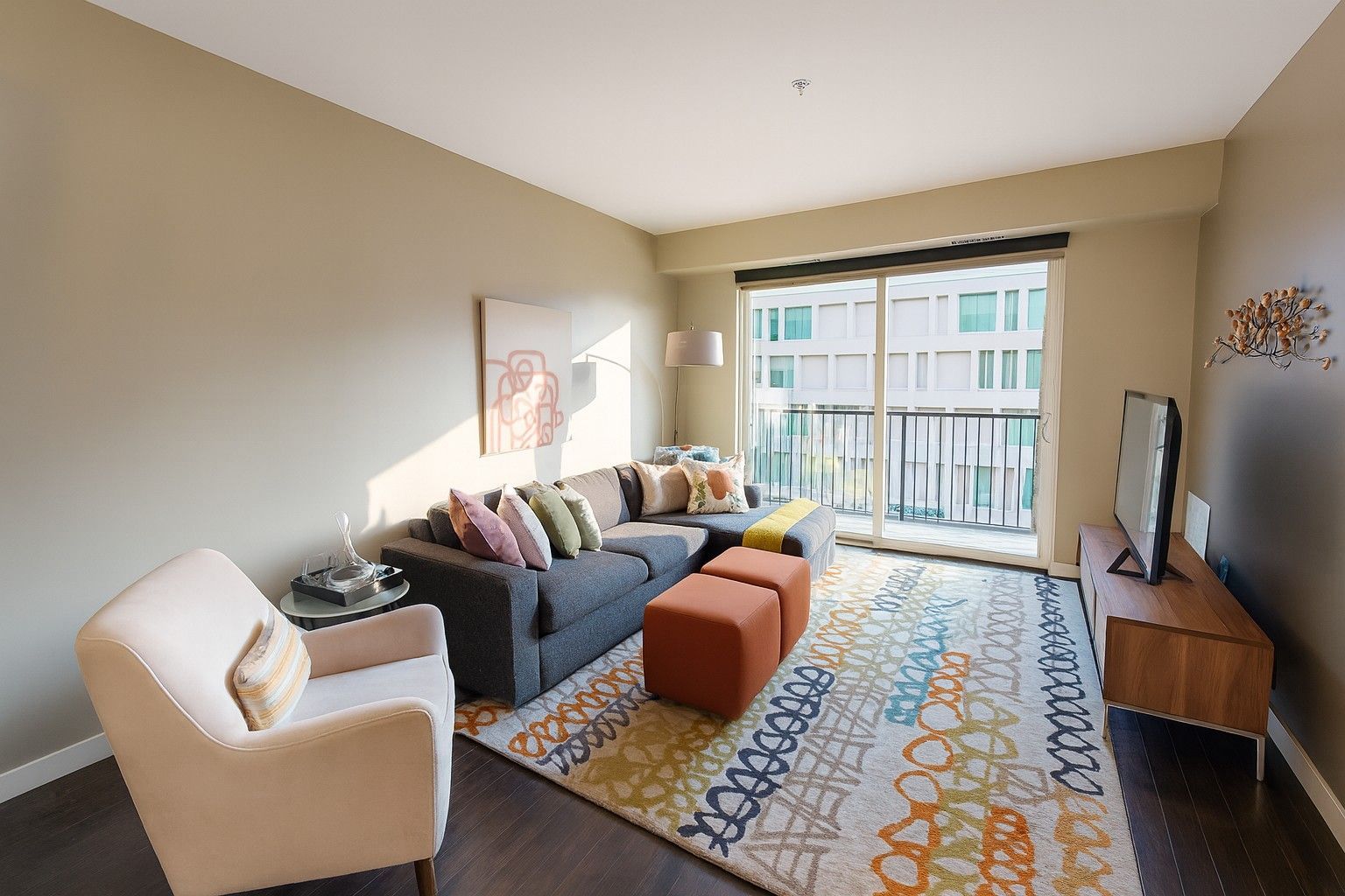 Bright living room with blue sofa, white chair, orange ottoman, TV, and sliding glass doors to a balcony.