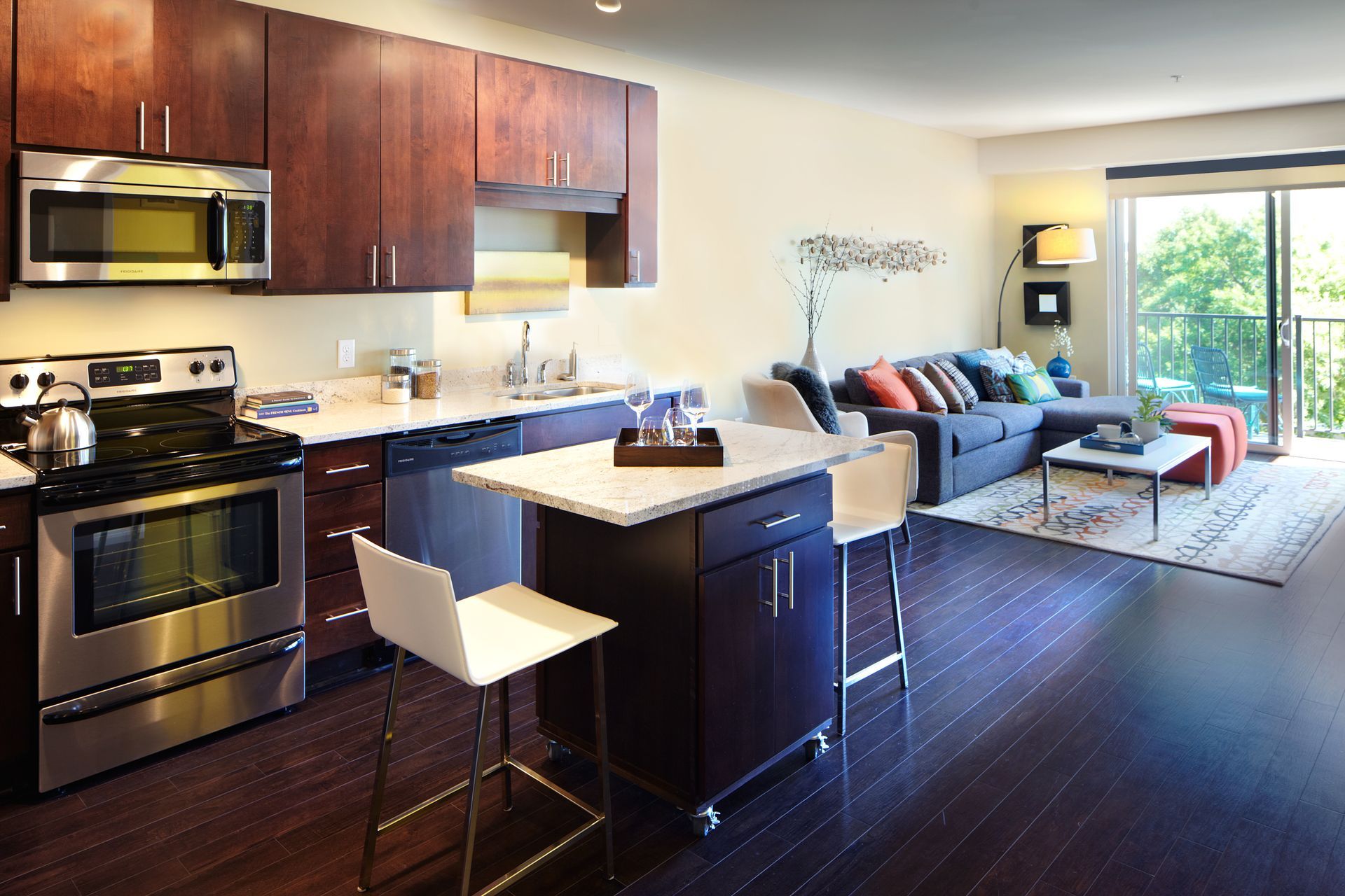 Modern open-plan apartment kitchen and living room with dark wood cabinets, island, sofa, and balcony view