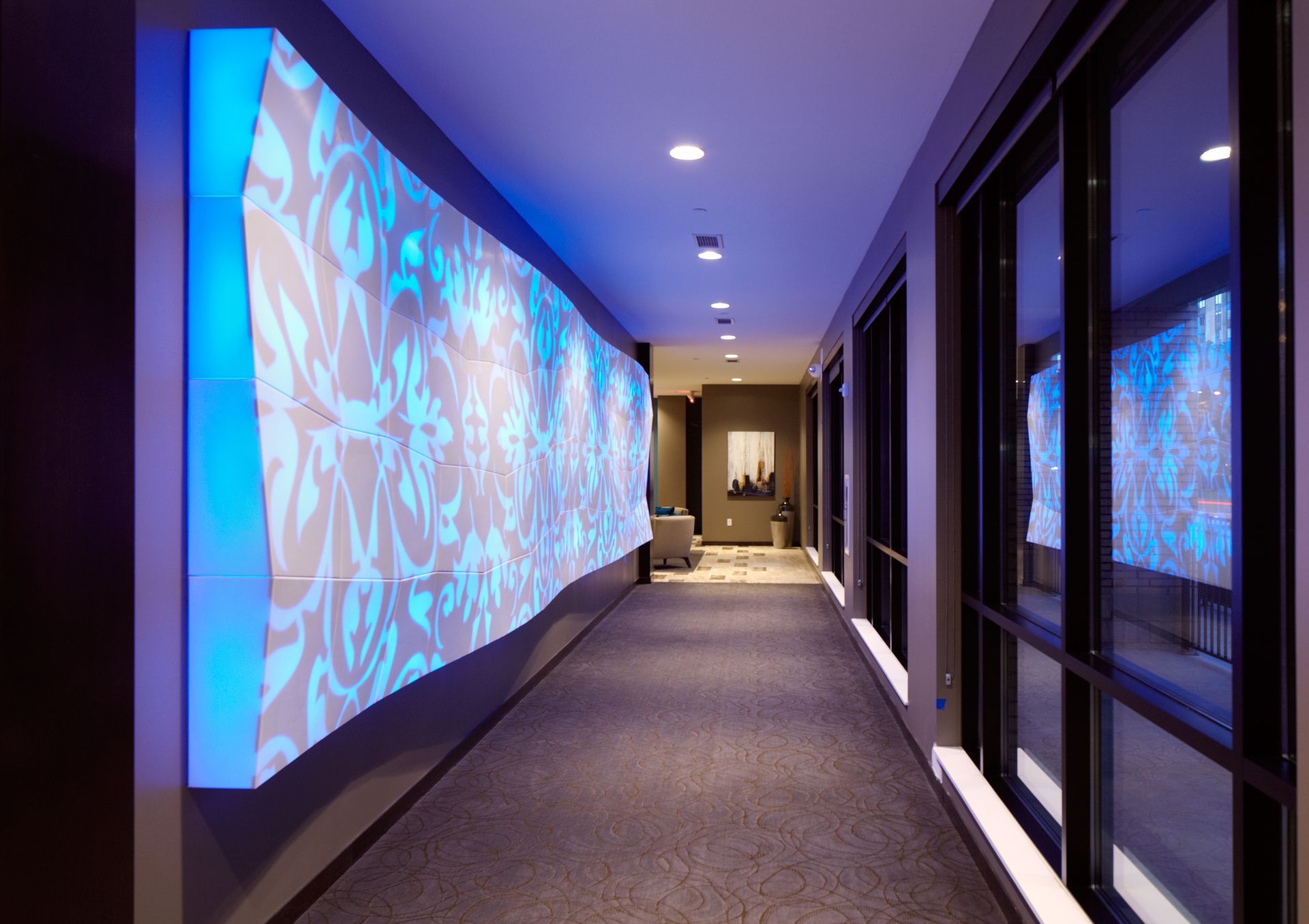 Modern hallway with blue patterned wall projections, carpeted floor, and glass panels on the right