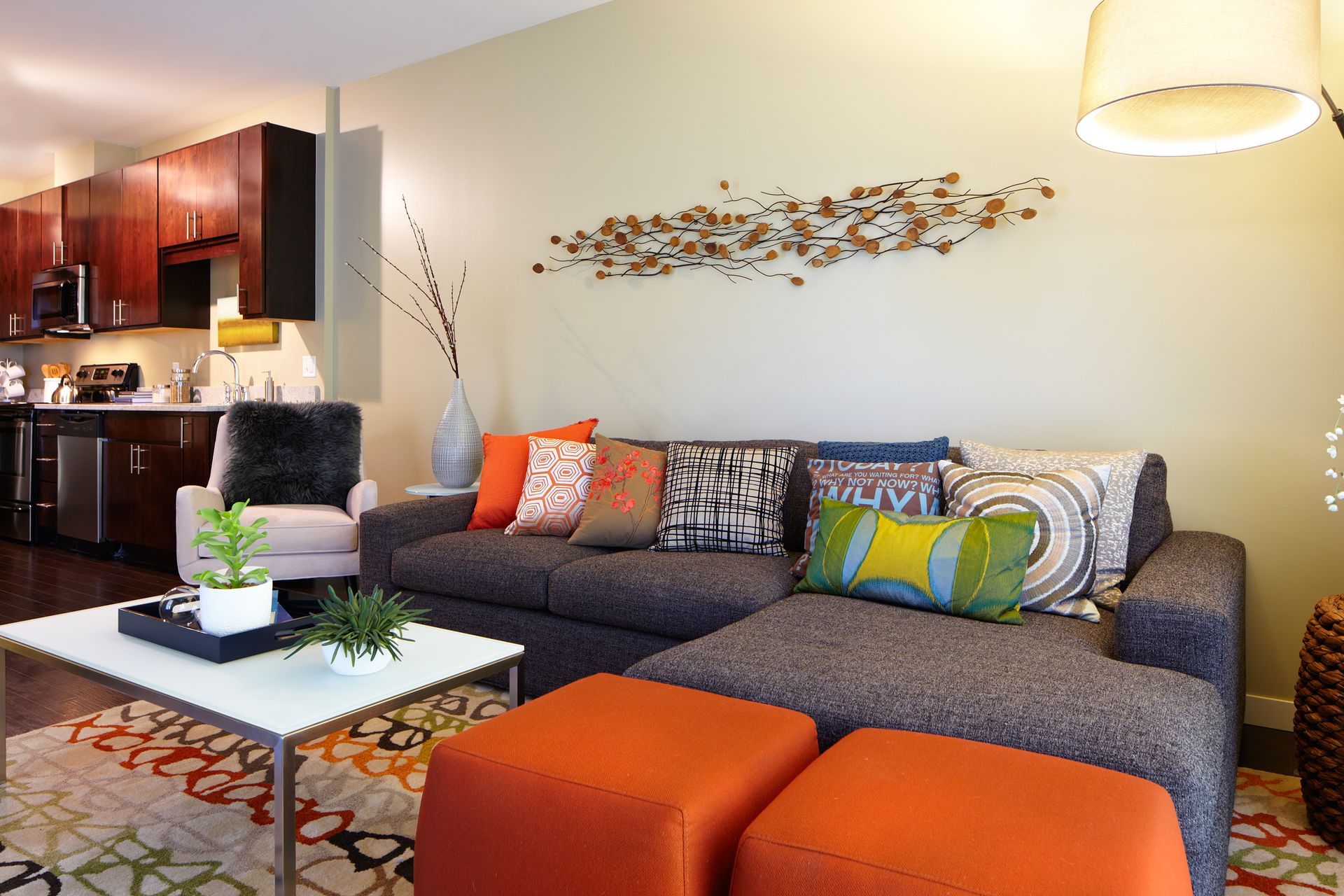 Modern living room with gray sectional, orange ottomans, white coffee table, and open kitchen.