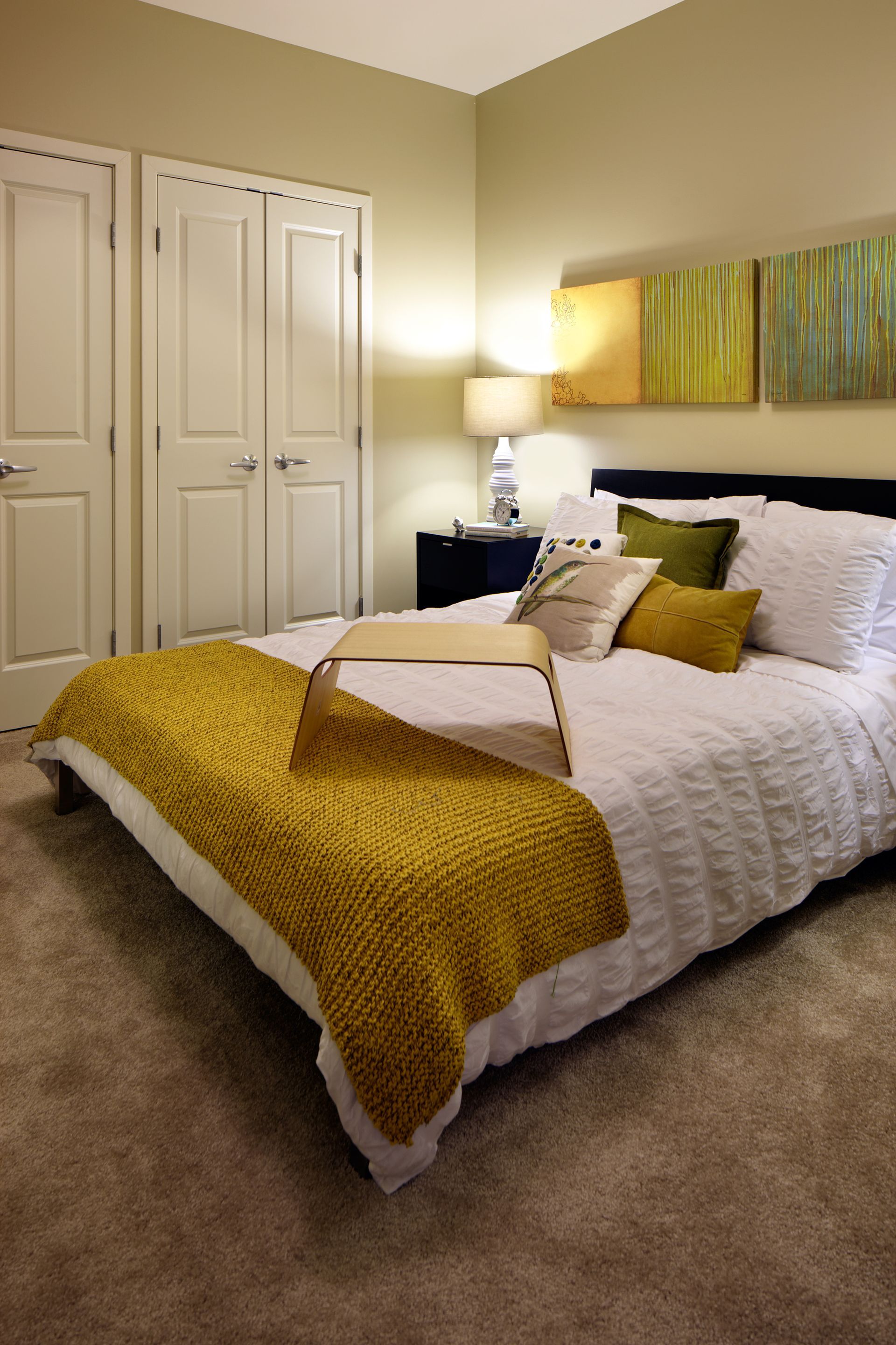 Cozy bedroom with a white bed, mustard throw blanket, lamp, closet doors, and abstract wall art.
