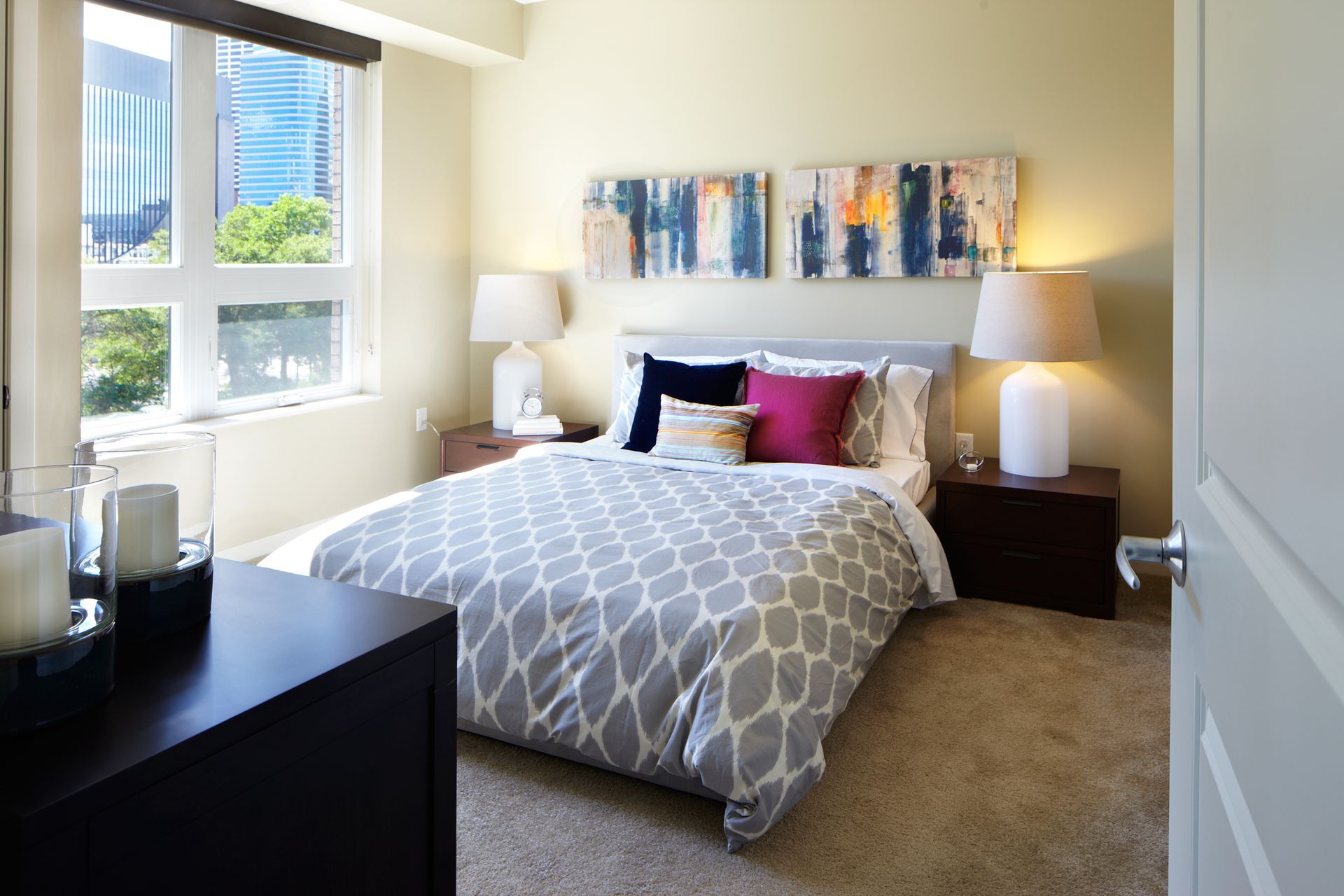 Bright bedroom with a bed, bedside lamps, colorful wall art, and a window with city view.