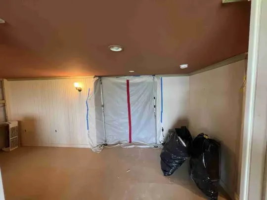 Empty room with white plastic sheeting over a wall, brown ceiling, and black trash bags on the floor