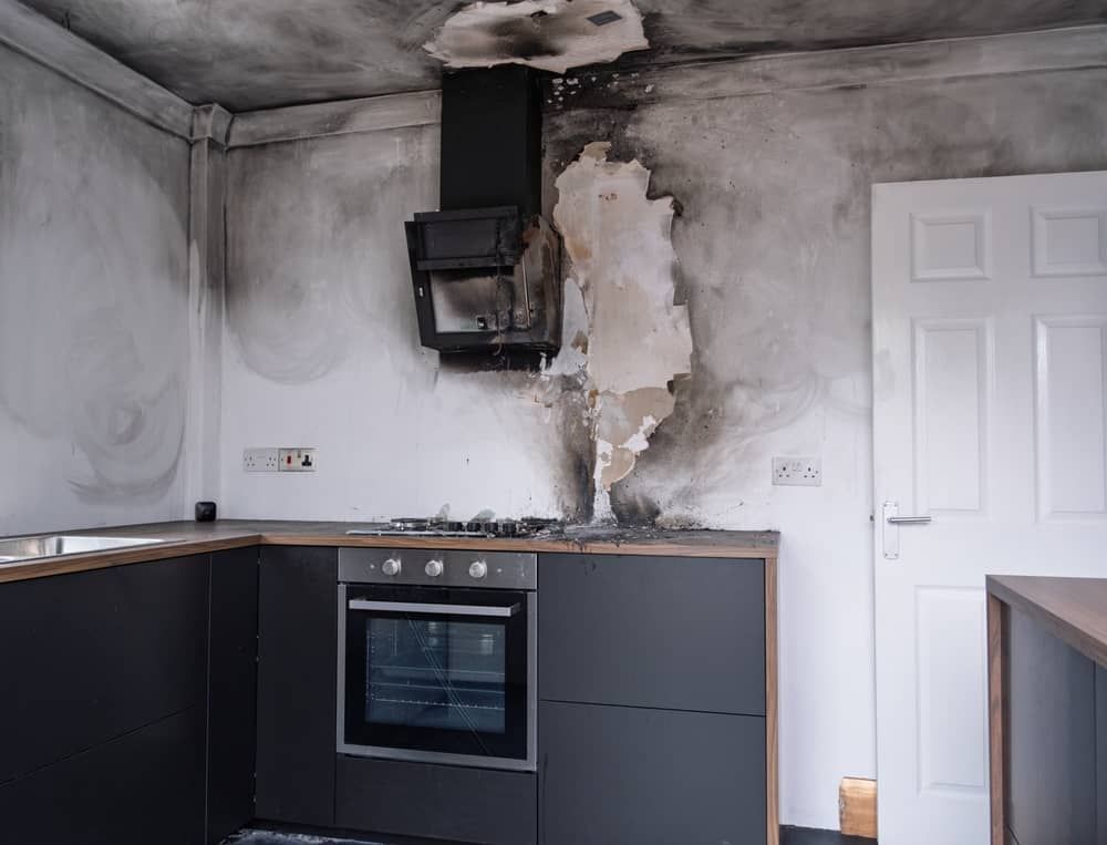 Damaged kitchen wall with a black oven and torn plaster around the range hood, beside white cabinets and a door
