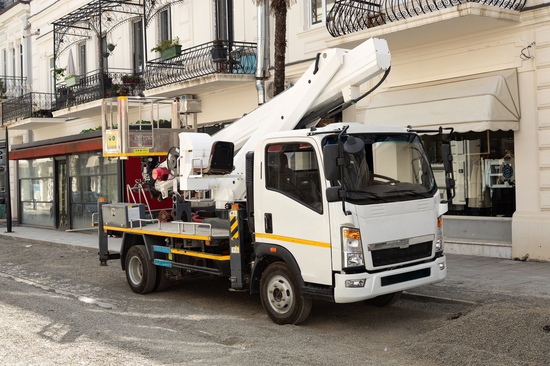 White utility truck with a raised boom parked on a street beside storefronts and apartment balconies White utility truck with a raised boom parked on a street beside storefronts and apartment balconies
