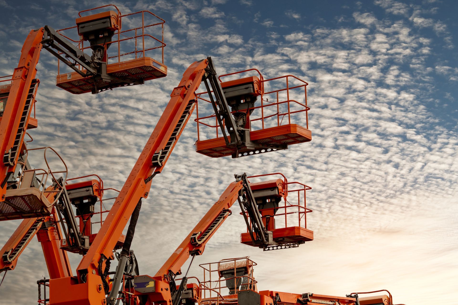 Orange truck crane rental units with raised hydraulic booms against a cloudy sky.
