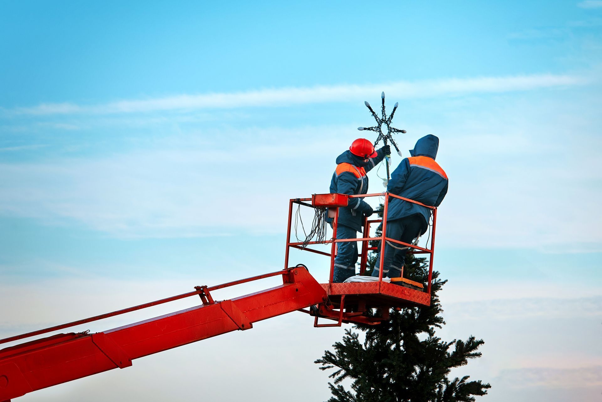 Two workers in orange safety gear on a red cherry picker trimming a tall tree against a blue sky