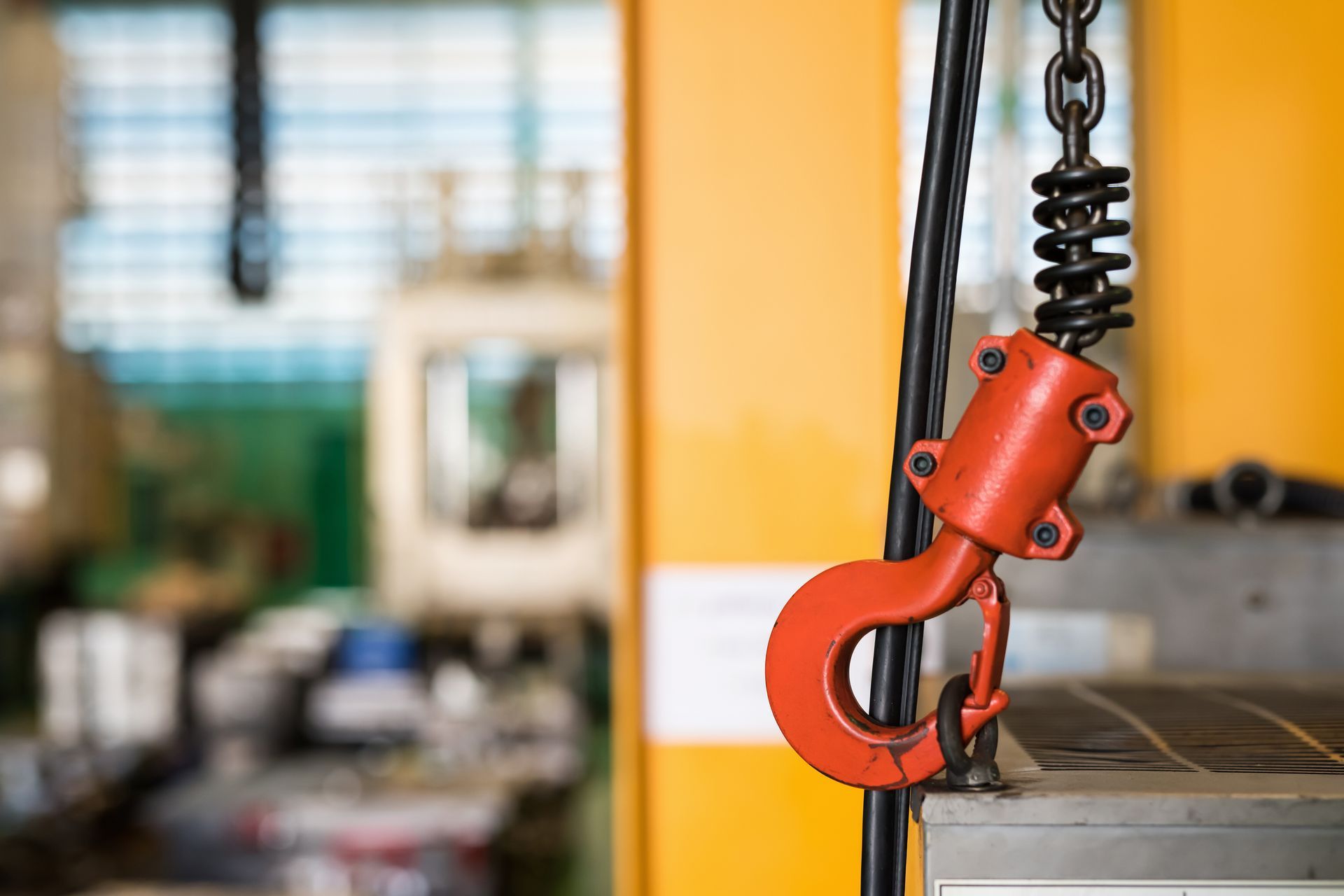 Orange industrial hook hanging from a chain in a workshop, with a blurred workshop background. Orange industrial hook hanging from a chain in a workshop, with a blurred workshop background.