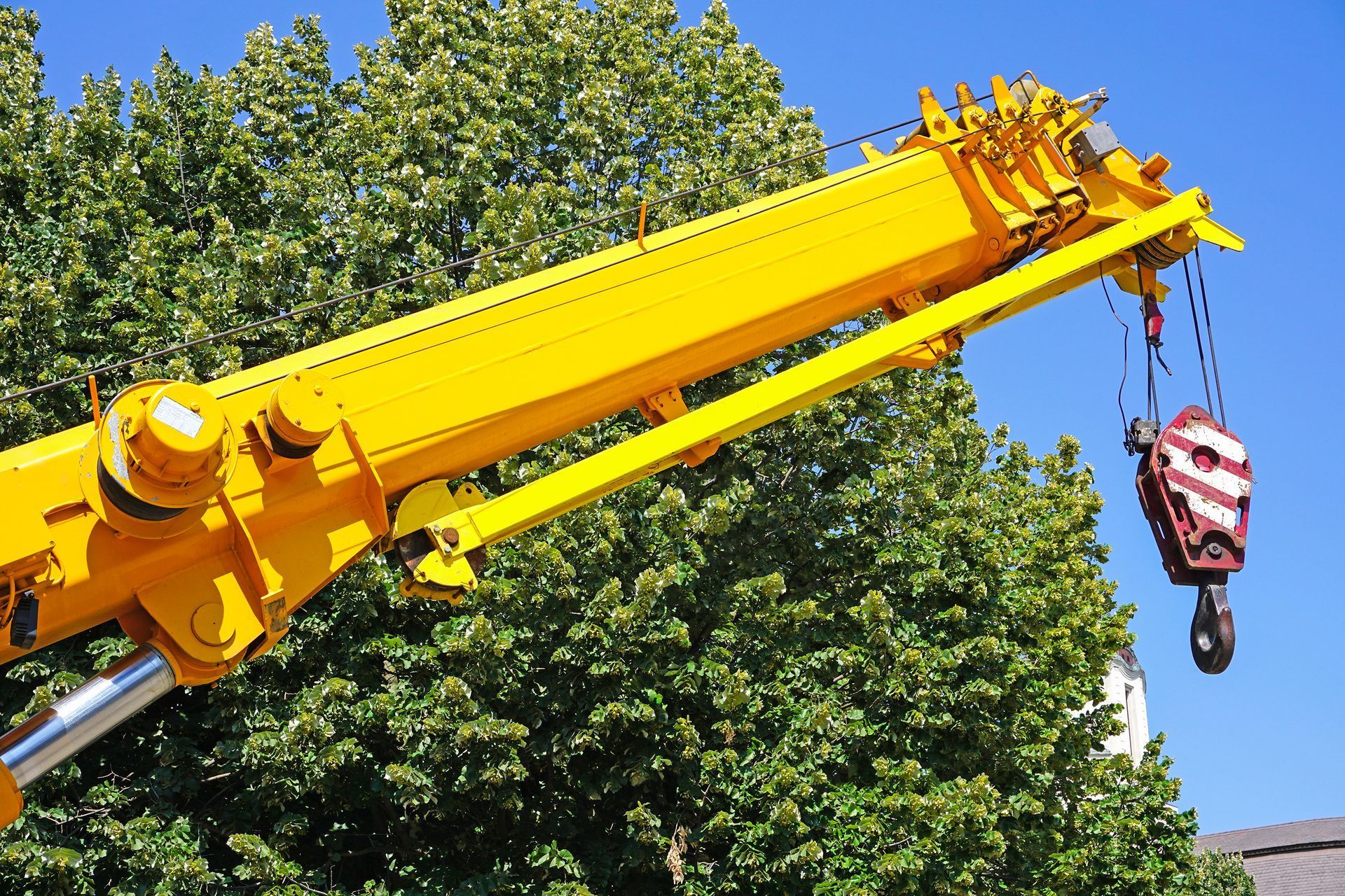 Yellow crane boom lifting a red-and-white hook block against green trees and blue sky