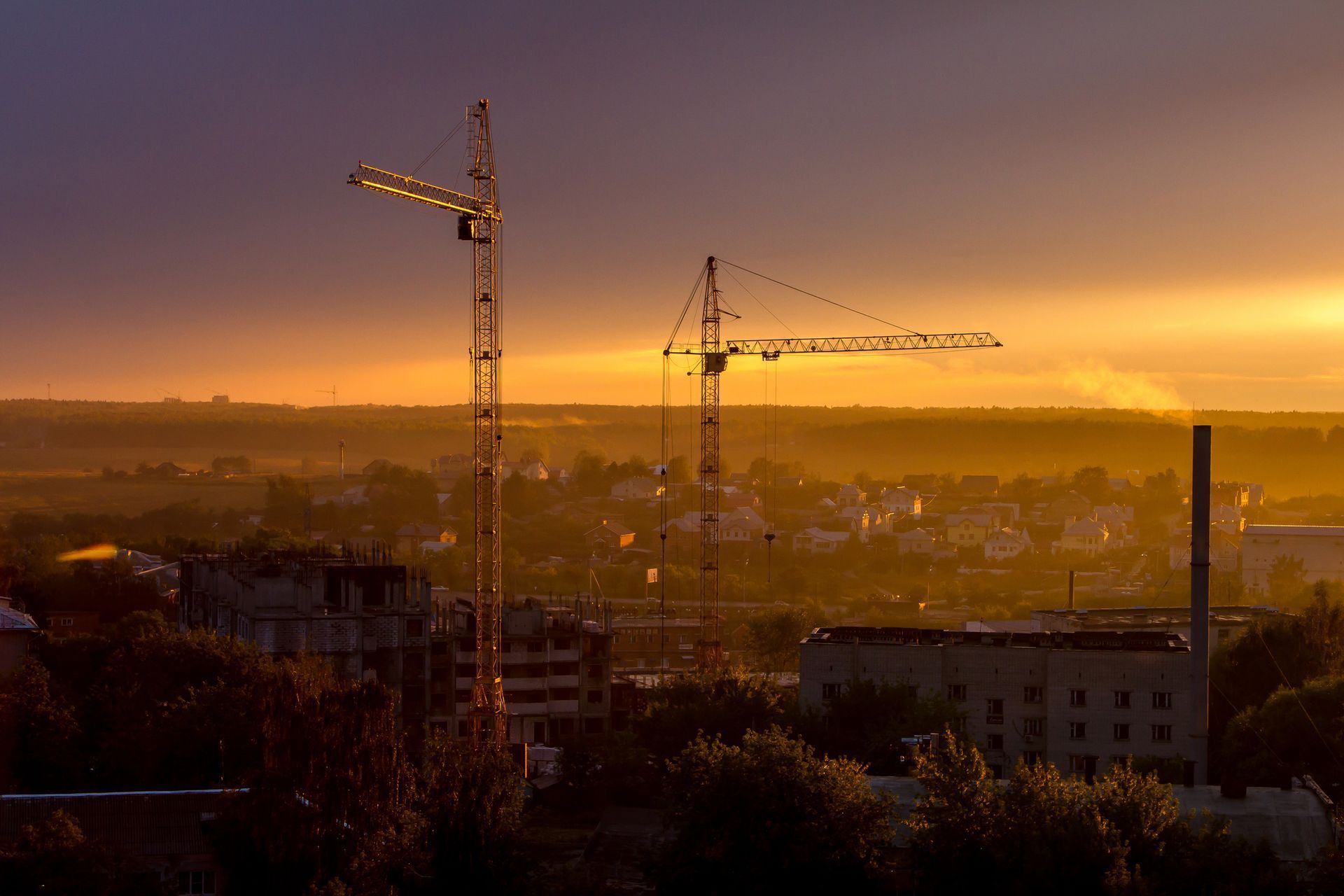 Sunset over a city skyline with two construction cranes silhouetted against a golden sky