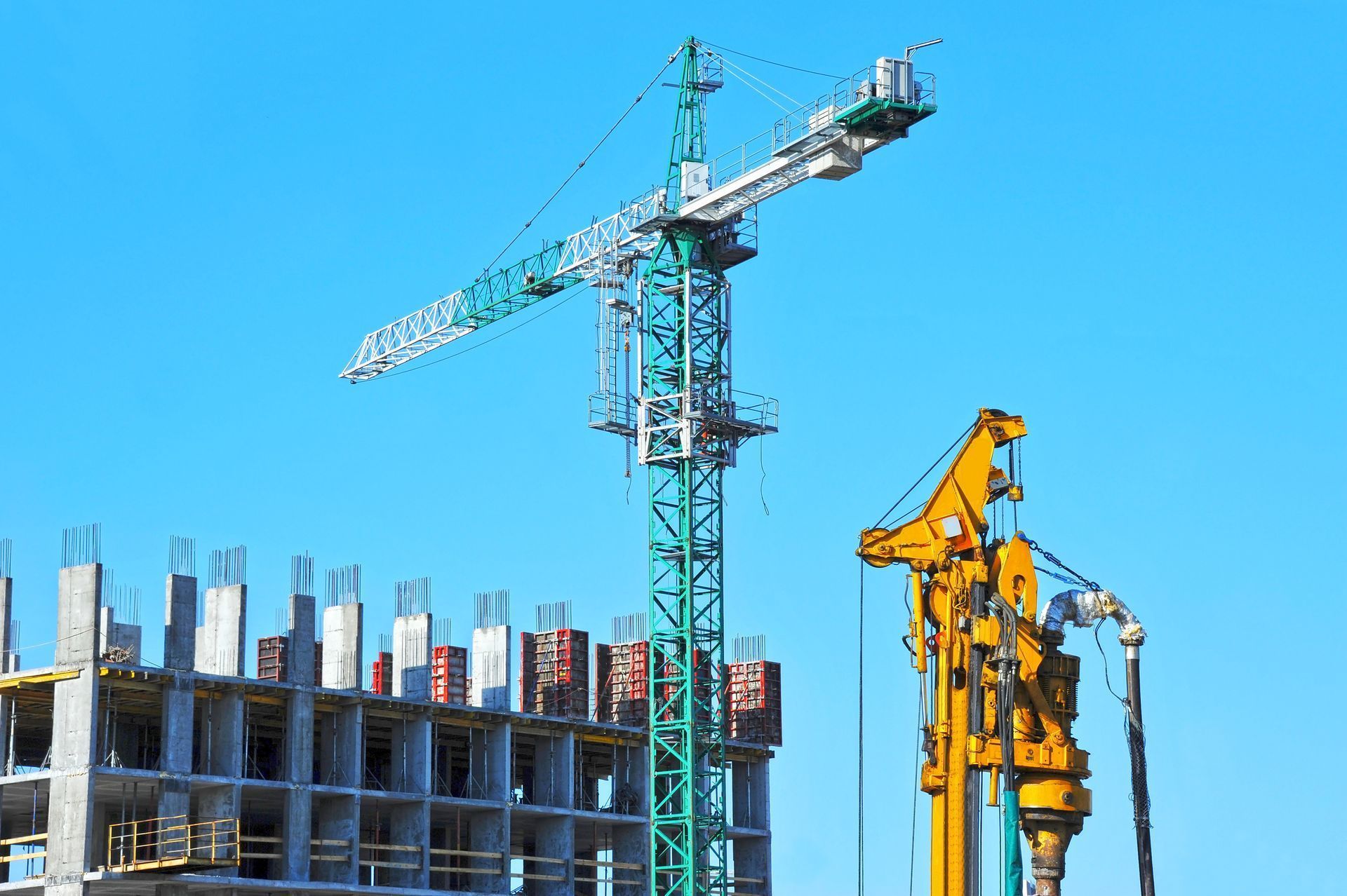 Construction site with a tall green crane, yellow machinery, and a partially built concrete structure against blue sky Construction site with a tall green crane, yellow machinery, and a partially built concrete structure against blue sky