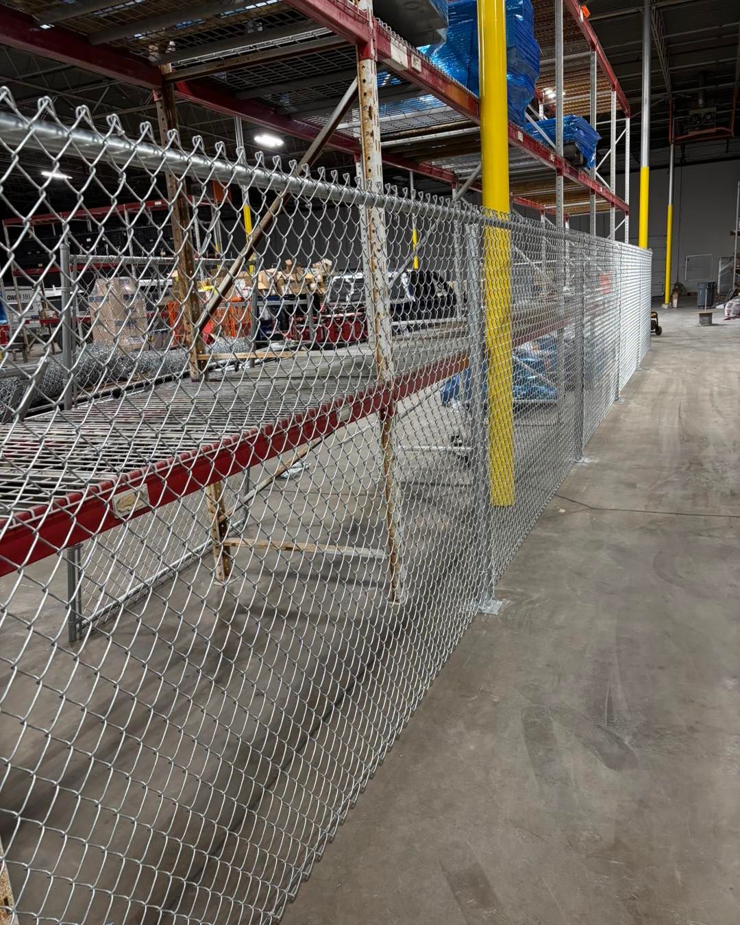 Chain-link fence separating an industrial warehouse aisle, with shelving and concrete floor.