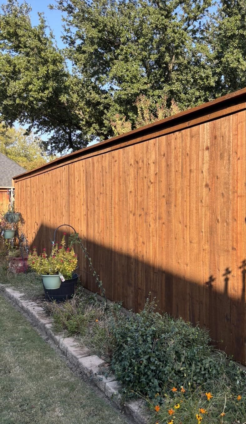 Brown wooden fence in a backyard next to plants and a flower bed under a blue sky.