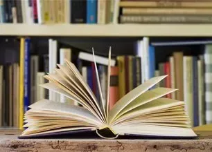 An open book sits on a wooden surface in front of a shelf filled with many colorful, upright books.