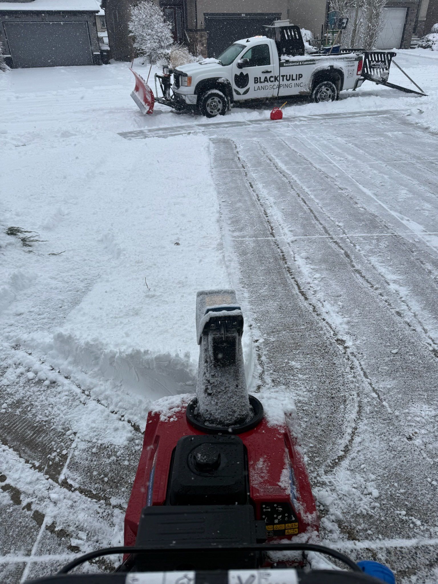 Snow blower clearing a snowy driveway, with a snow plow truck in the background.
