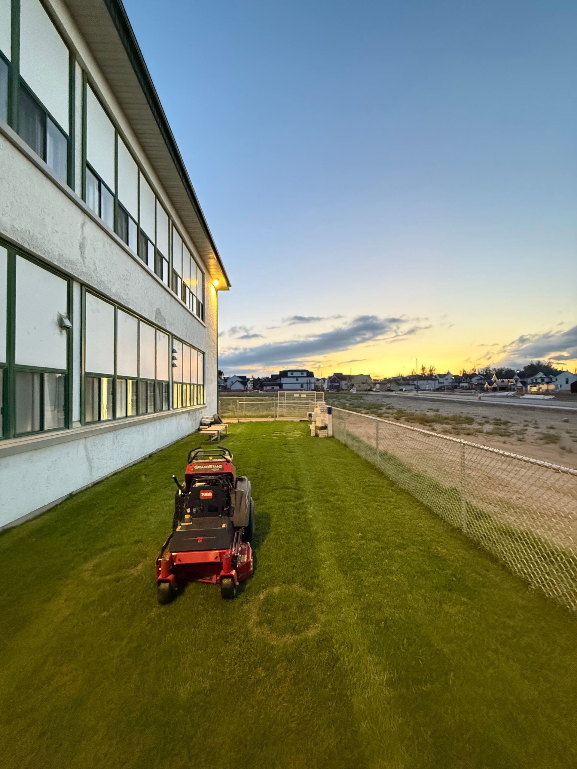 Red lawn mower parked by a white building beside a gravel lot at sunset